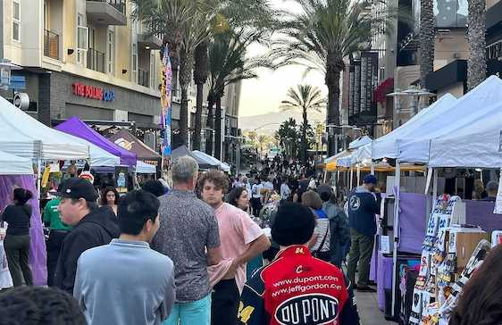 Crowded outdoor market with booths and people. Buildings and palm trees line the street.