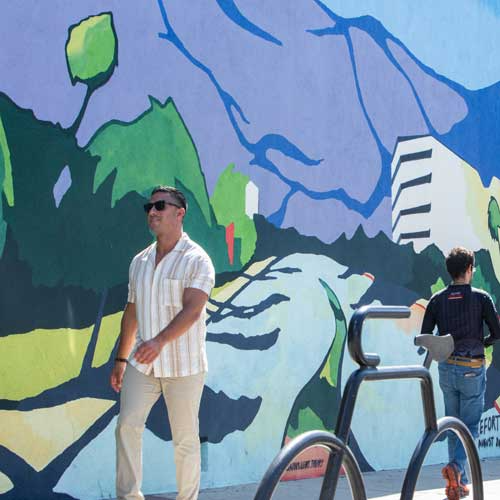 Man walking past a colorful mural. Mountains and buildings depicted. Another person nearby.