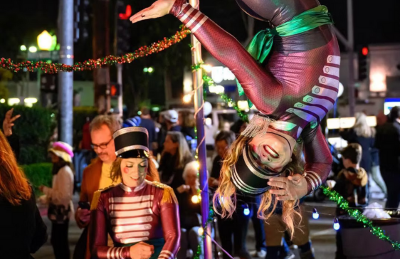 Street performer upside down on a pole, wearing body paint. Person in marching band uniform watches in a crowd.