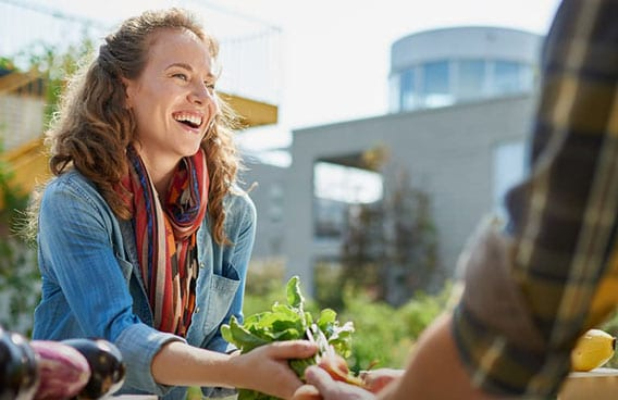 Woman laughing, receiving fresh greens outdoors. Bright sunshine, buildings in the background.