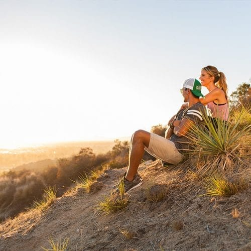 Couple sitting on a hillside, looking at a sunset; man in hat, woman has arm around him.