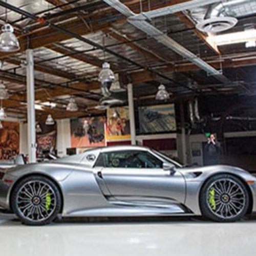 Silver Porsche 918 Spyder parked in a well-lit garage, featuring green brake calipers and artwork on the walls.