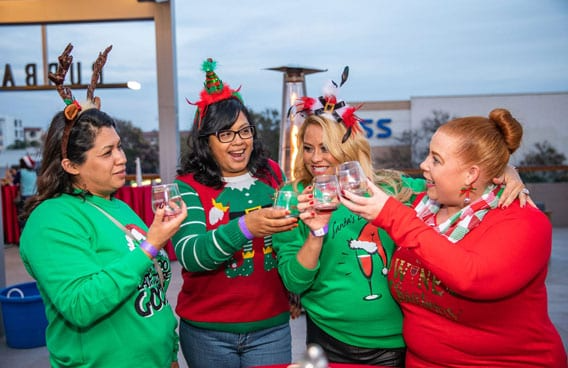 Four women in festive sweaters toast with drinks outdoors, celebrating Christmas.