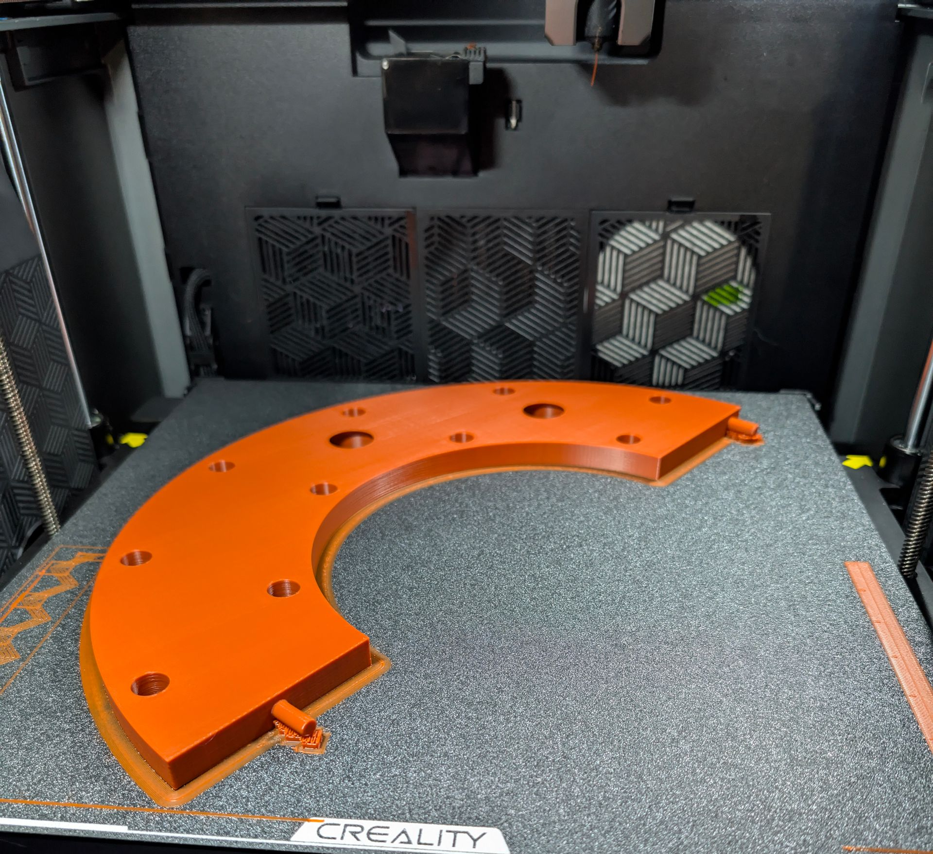 An orange, semi-circular 3D-printed part in a 3D printer, with ventilation and 