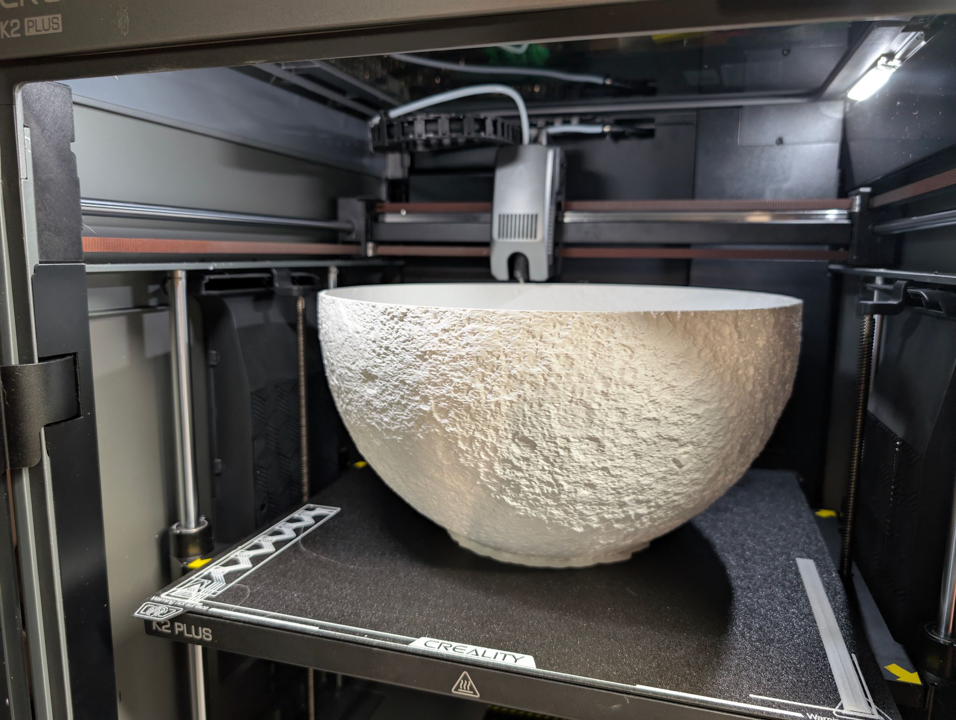 3D printer creating a large, moon. The machine is black with silver details, located in a workshop.