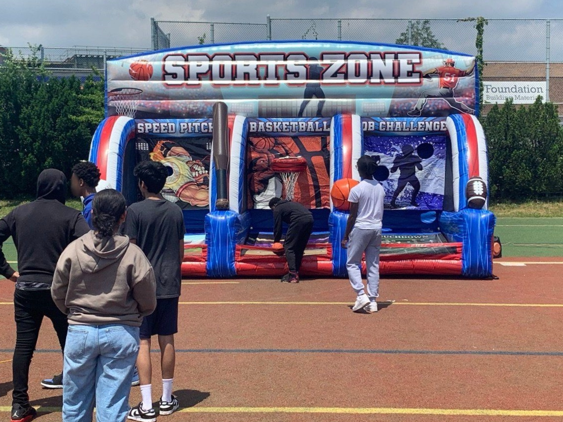 A group of people are standing in front of Sports Zone – an inflatable sports game: Speed Pitch (Baseball), Basketball, QB Challenge (Football)