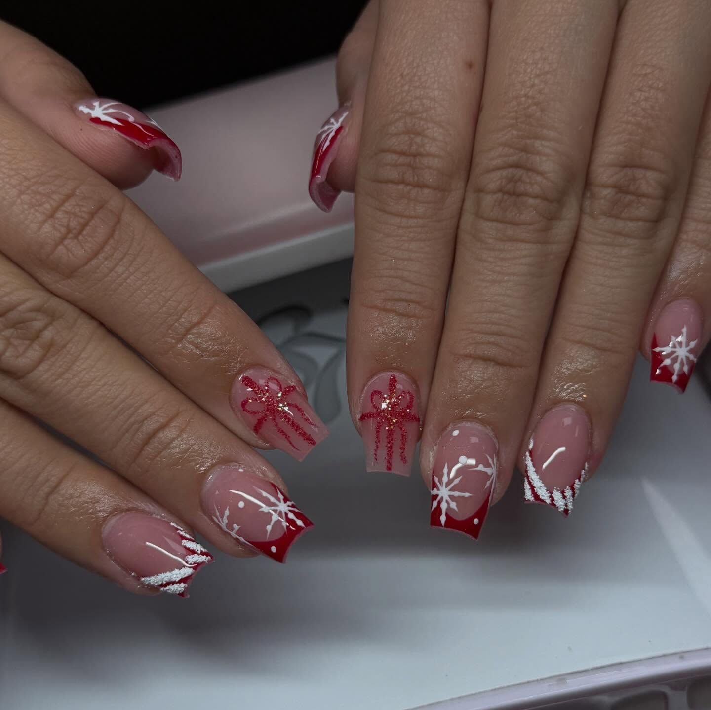 Hands with Christmas-themed nail art: red tips, snowflake designs, and gift bow accents.