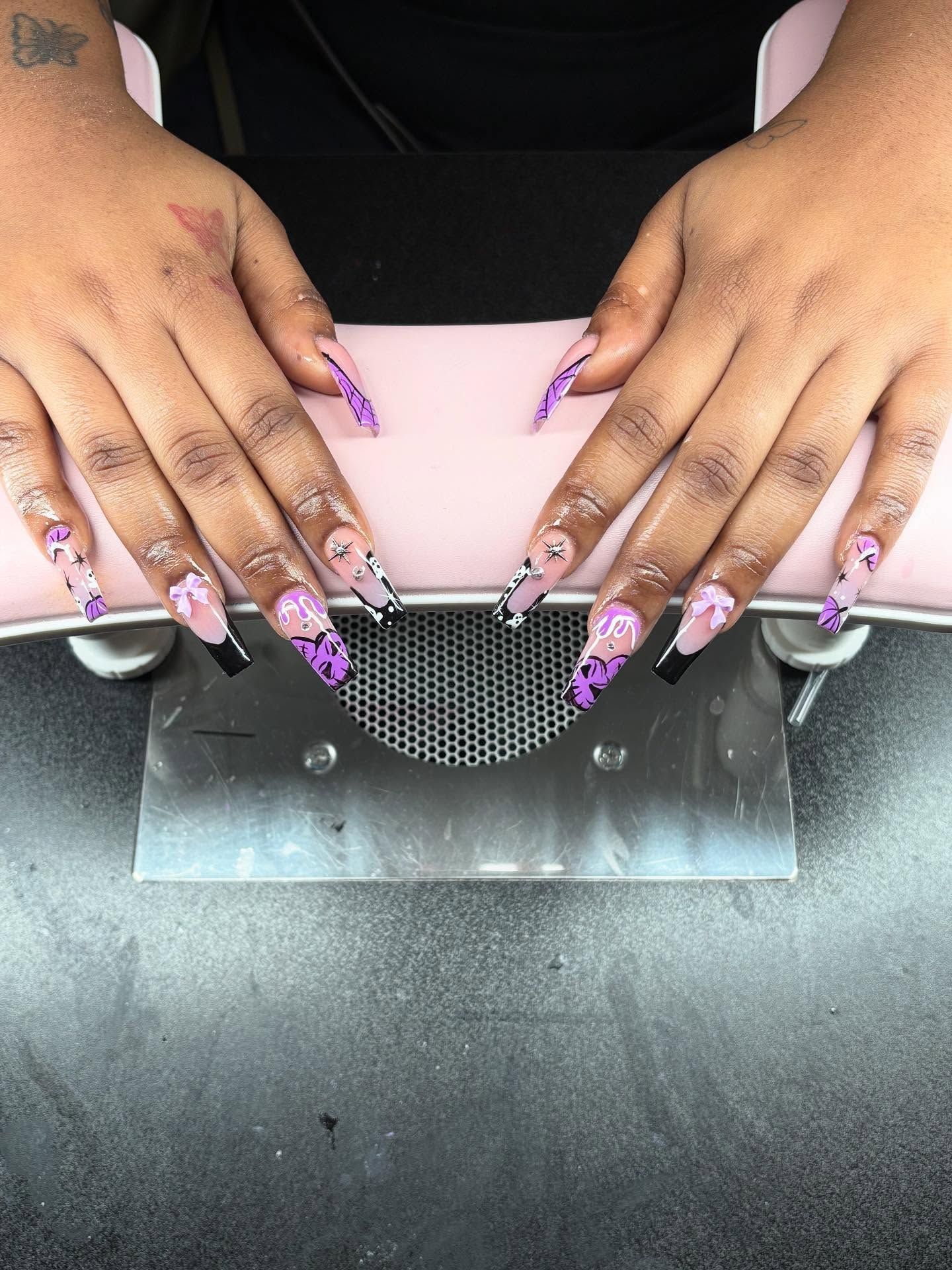 Hands with long, painted nails in shades of black, white, and purple, resting on a pink surface.