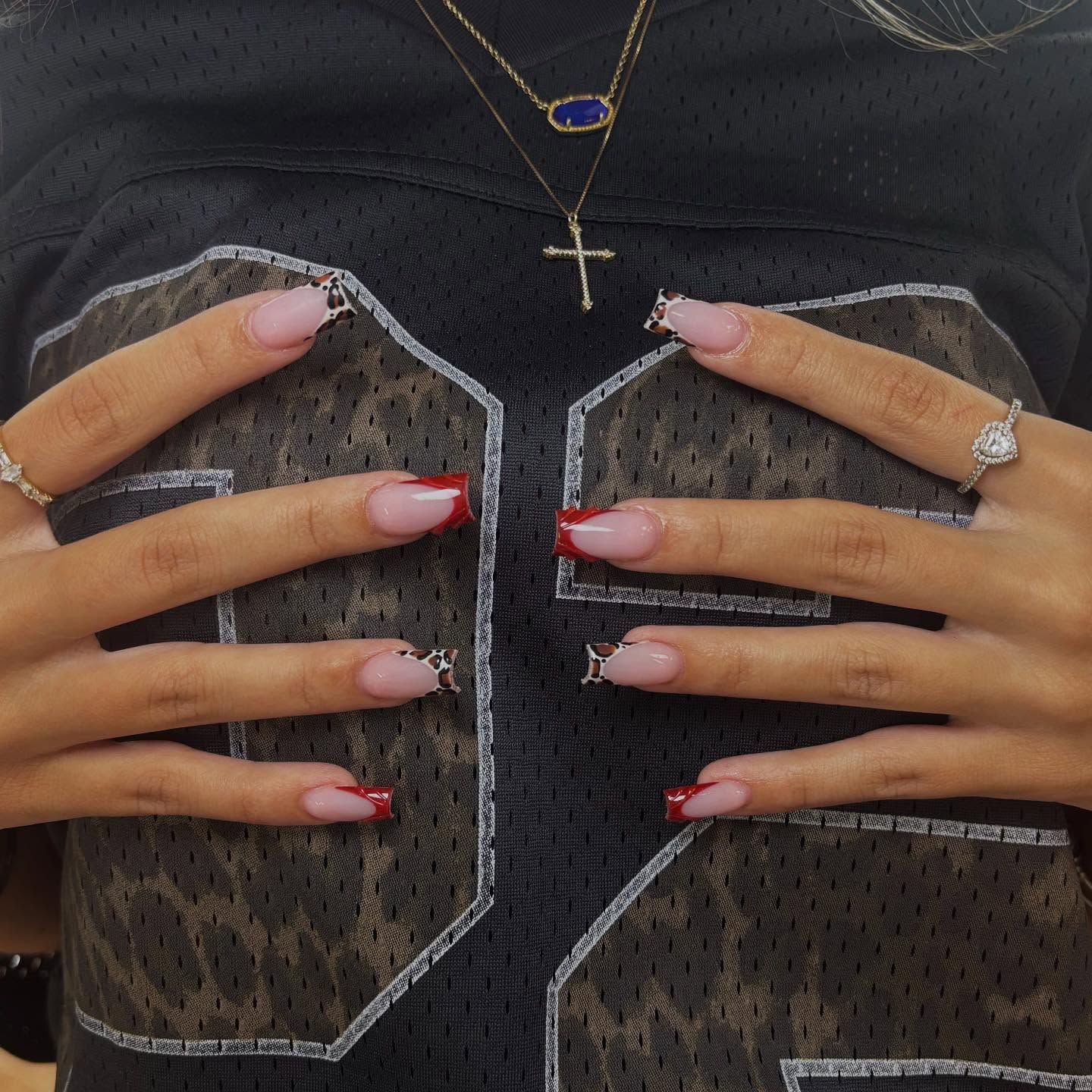 Hands with red-tipped nails on a black jersey with a leopard print pattern, wearing rings and necklaces.