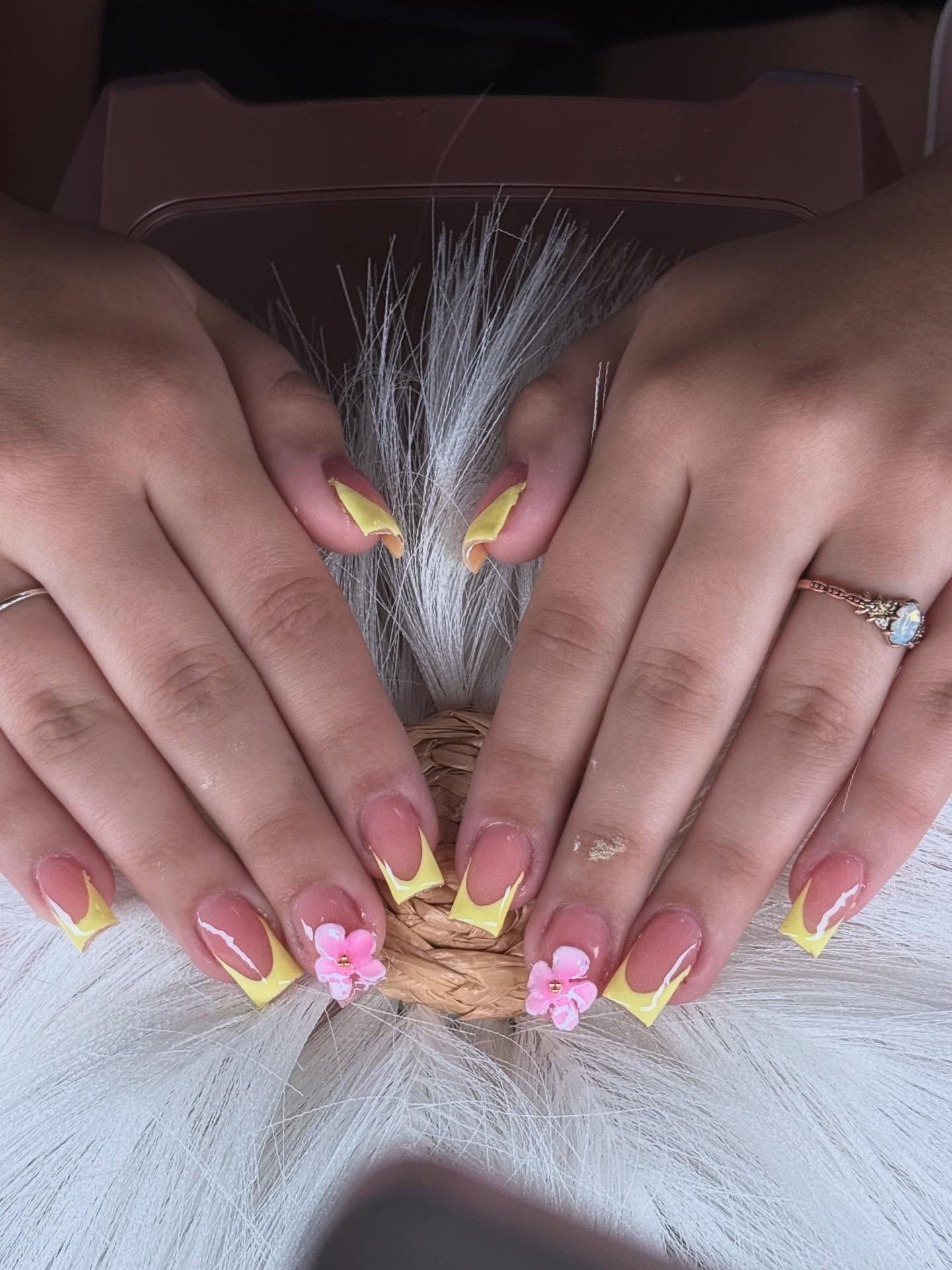 Hands with yellow French-tip acrylic nails, some with 3D pink flowers, resting on a white, feathery surface.