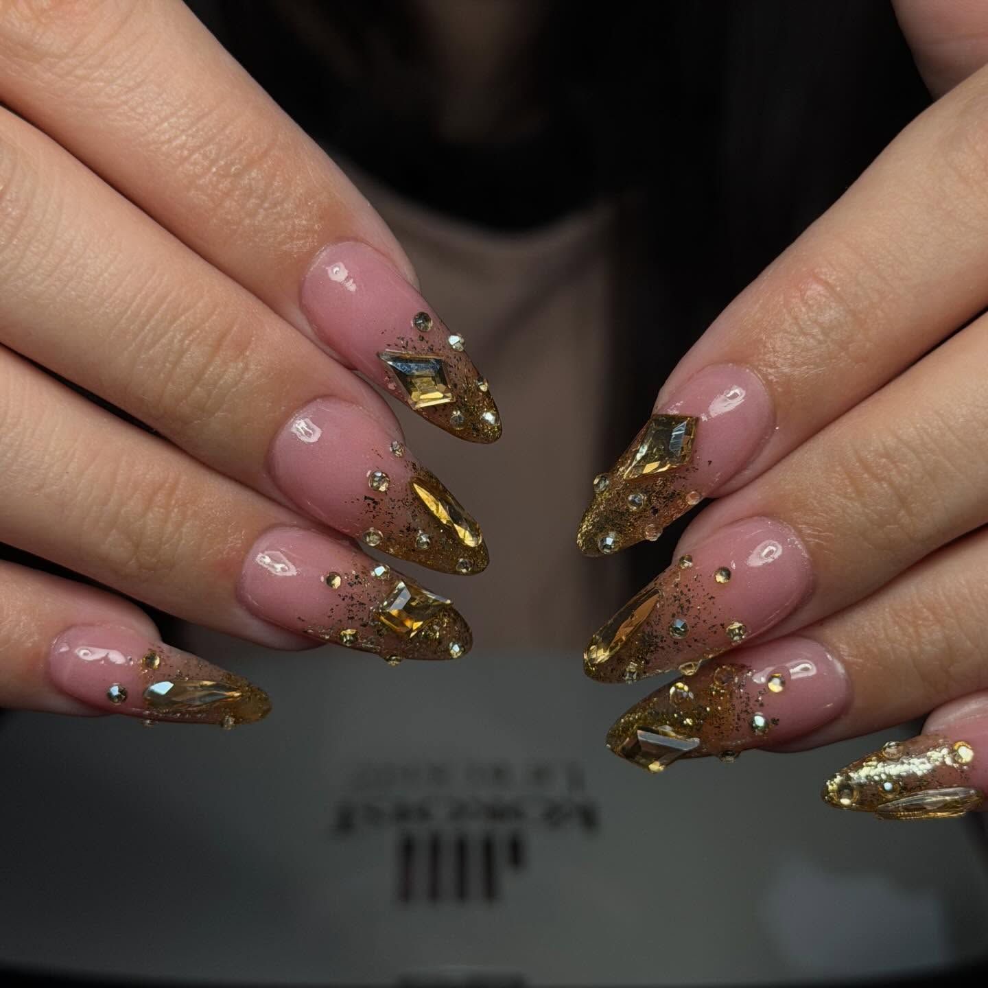 Close-up of hands with long, almond-shaped nails in nude and gold with gem embellishments.