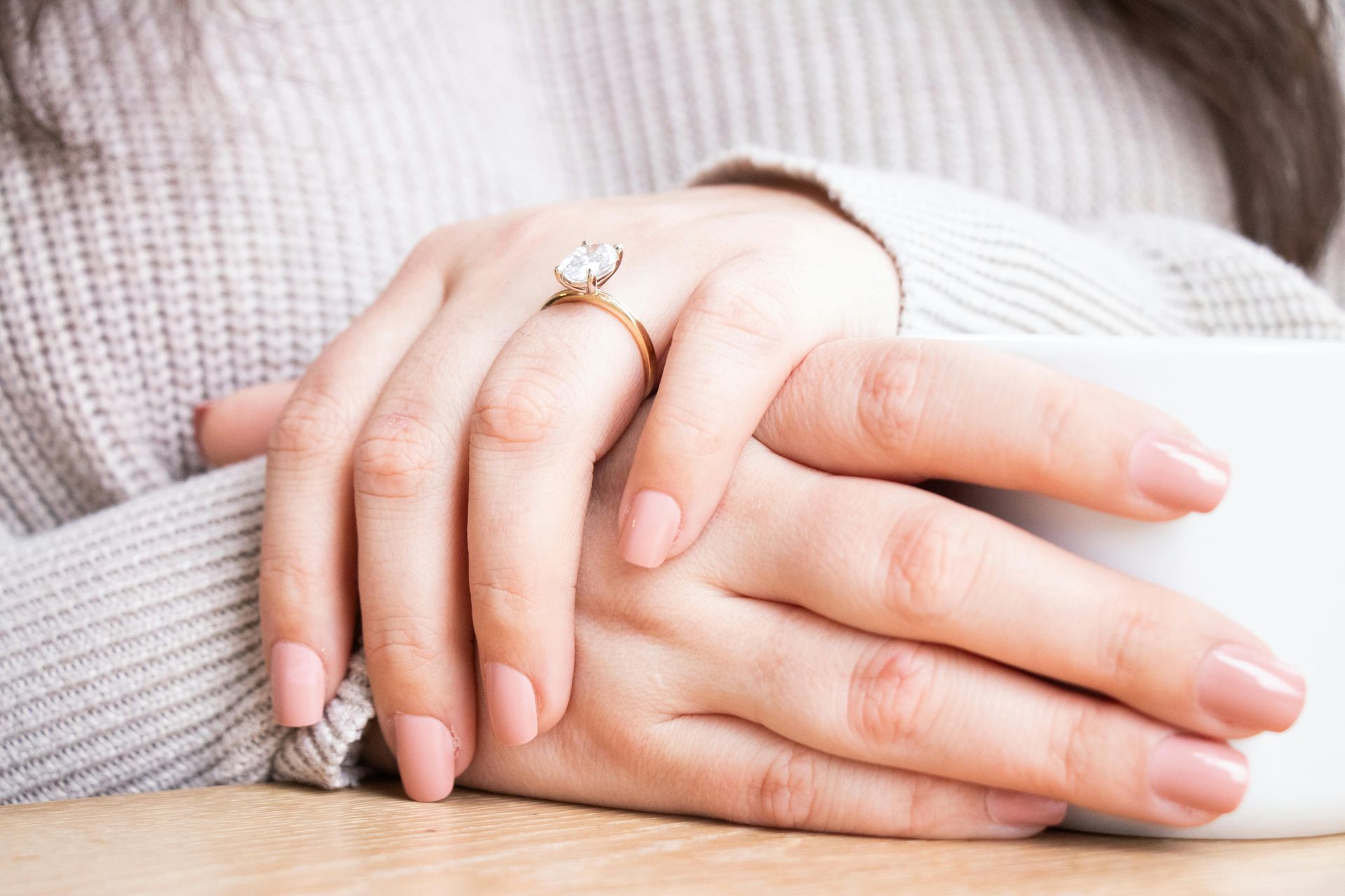 Hands with a diamond engagement ring, resting on a surface. Light pink nails.