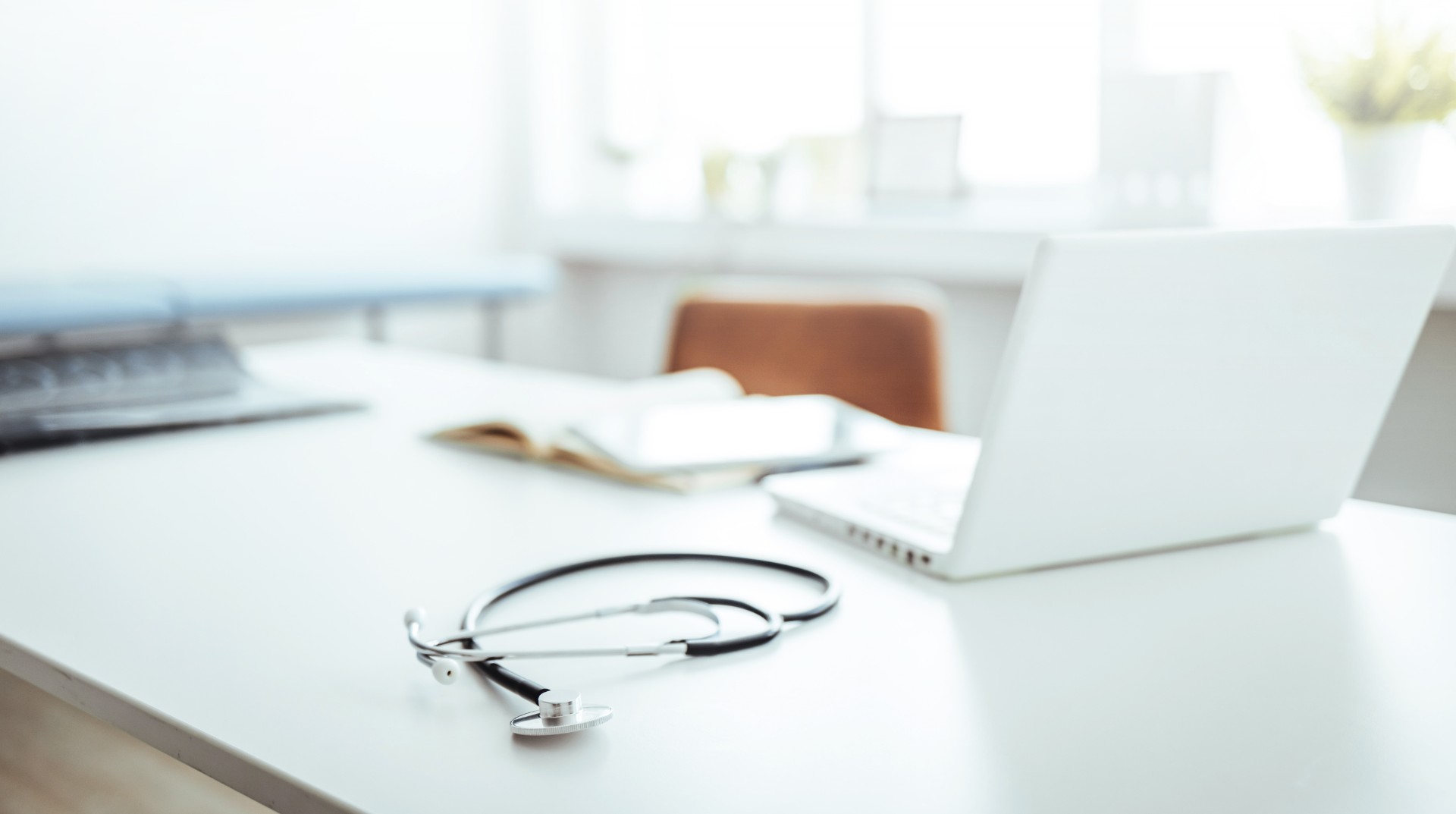 Stethoscope on a white desk in a doctor's office, with a laptop and open book.