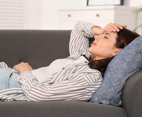 Woman lying on a sofa, hand on forehead, looking distressed. Gray sofa, blue pillow, neutral room.