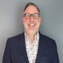 Man wearing glasses smiles in a navy blazer over a patterned shirt.