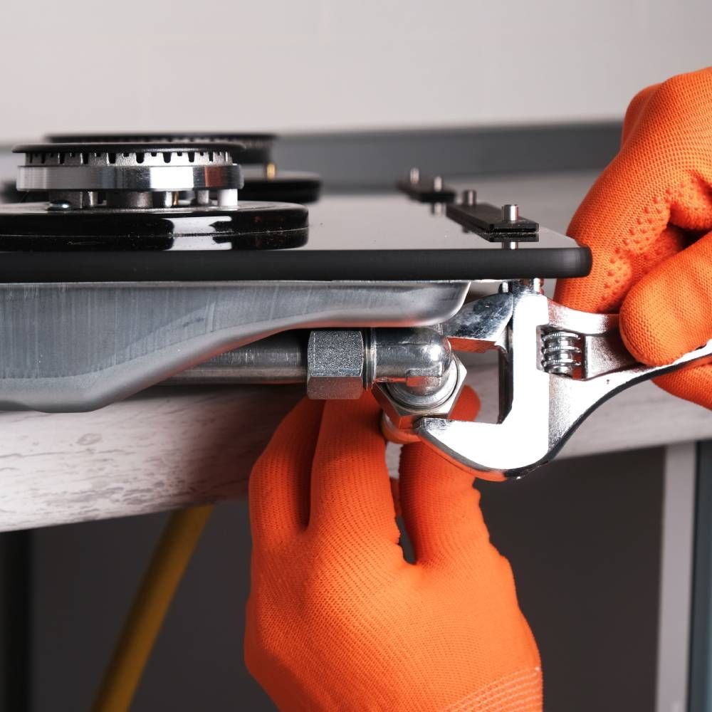 A person wearing orange gloves is fixing a stove with a wrench