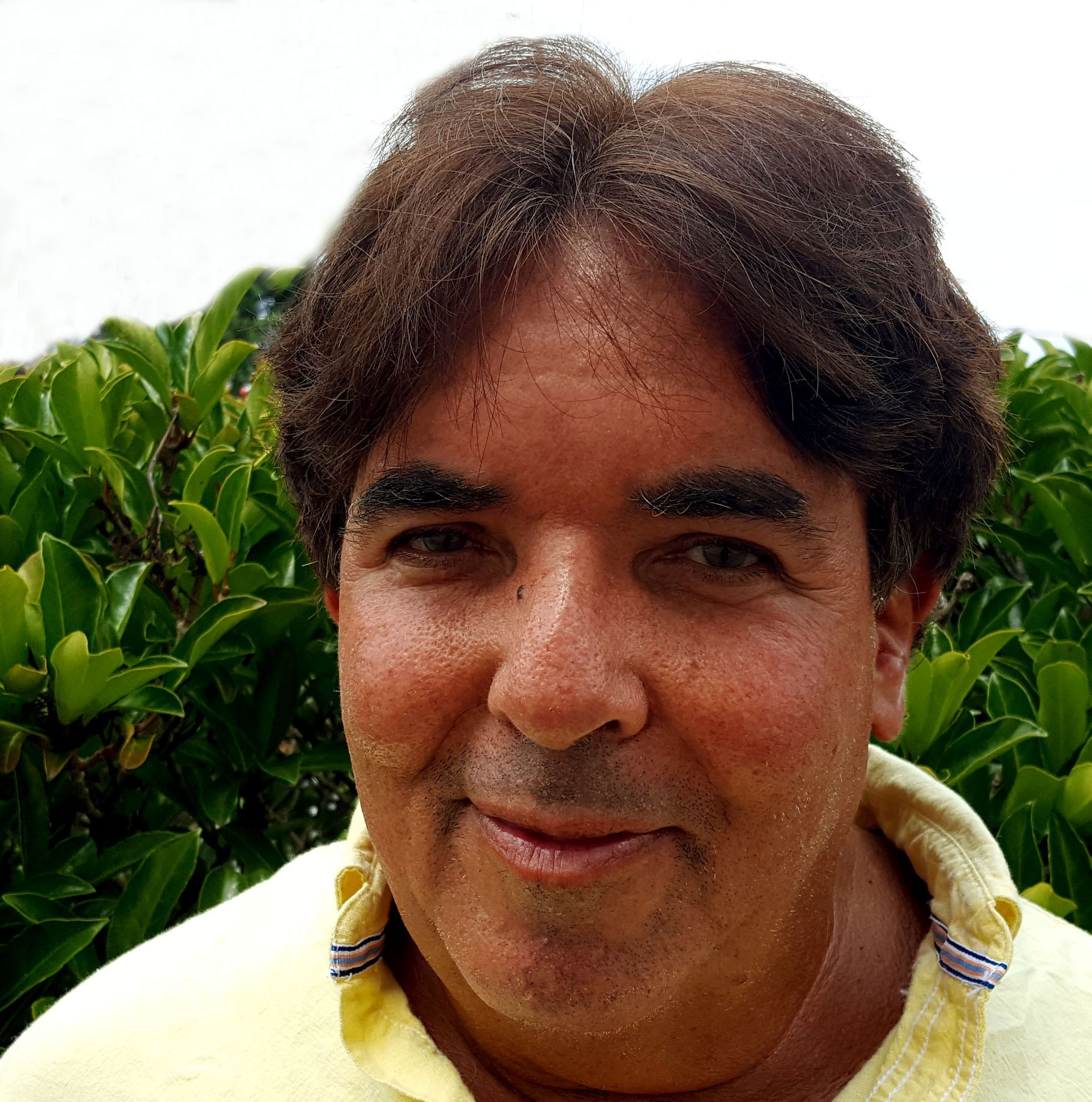 A man in a yellow shirt is smiling in front of a bush