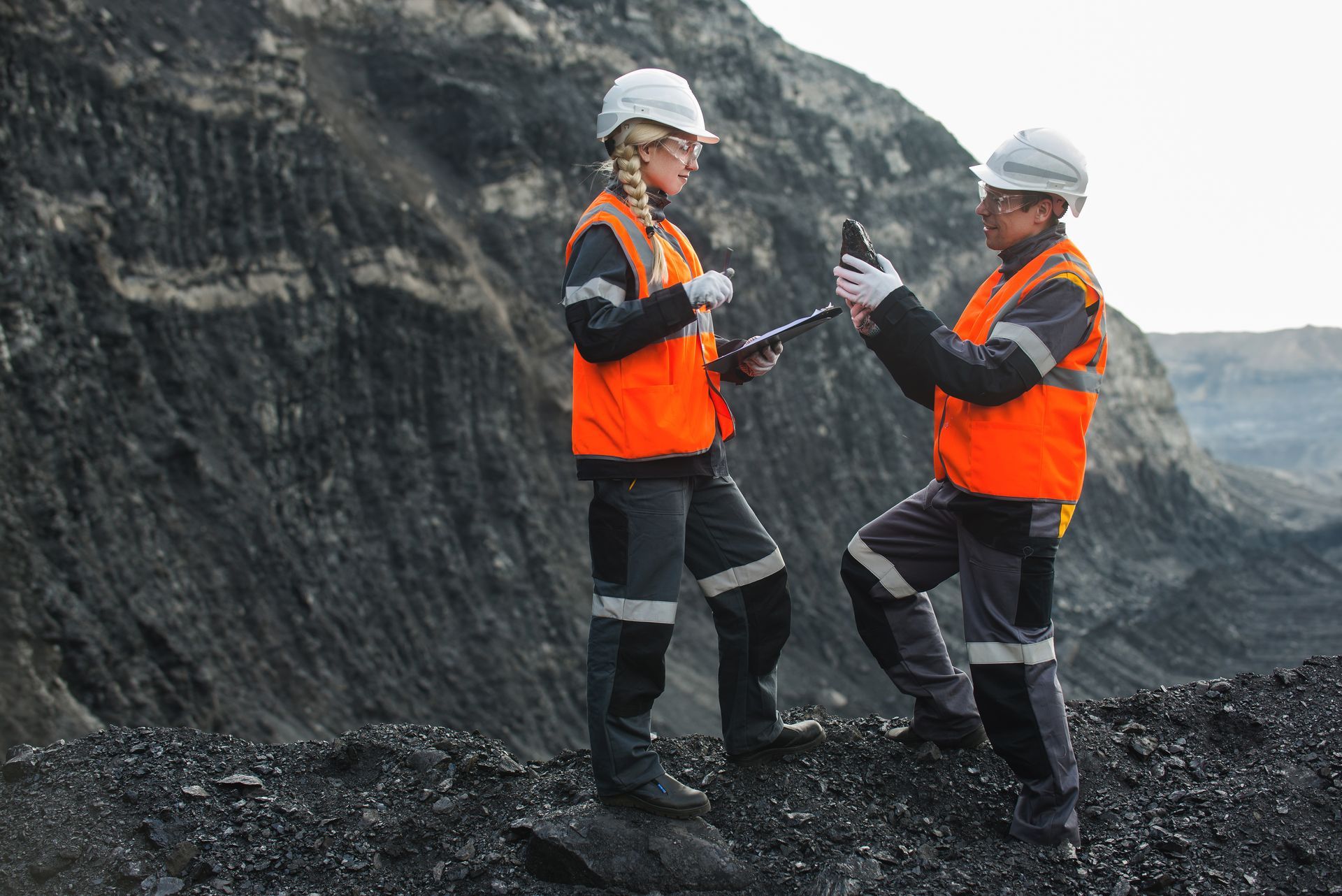 Two workers in orange safety vests and hard hats standing on a rocky quarry, one holding a clipboard.