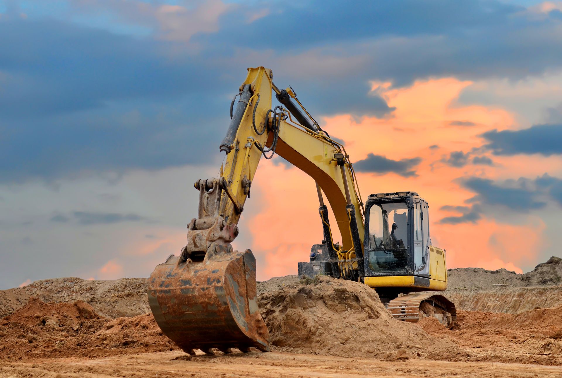 Yellow excavator digging dirt at a construction site under a cloudy sunset sky