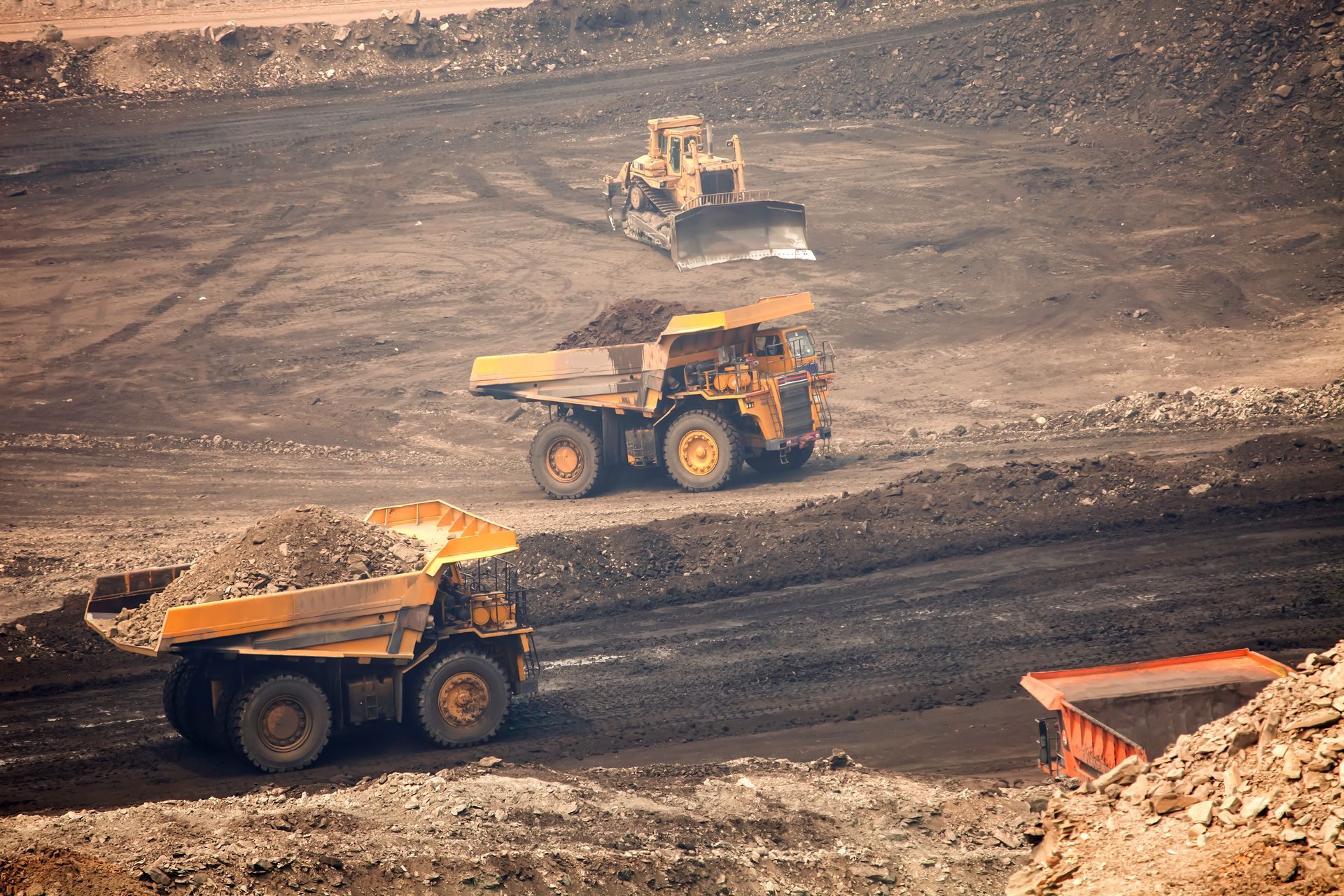 Three mining haul trucks on a dusty open-pit site, carrying earth and driving along terraced roads.