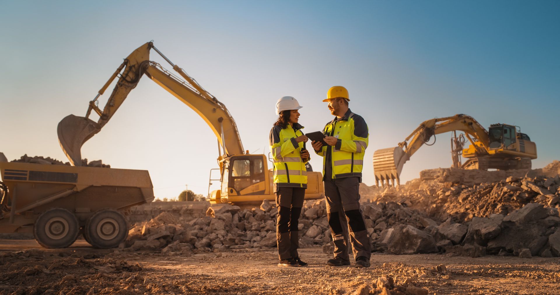 Two workers in safety gear stand at a construction site with excavators and dump trucks at sunset