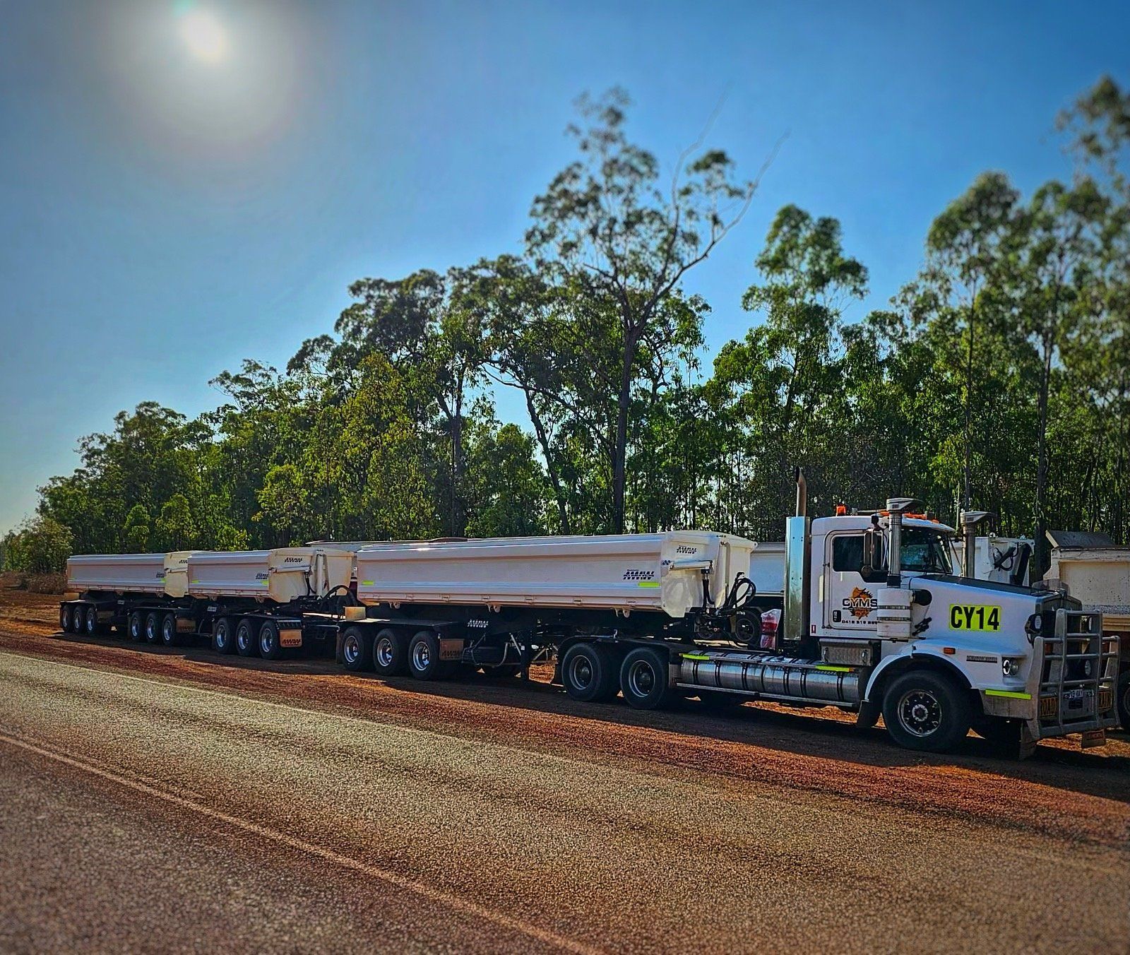 White tanker truck hauling trailers on a dirt road under a bright blue sky.