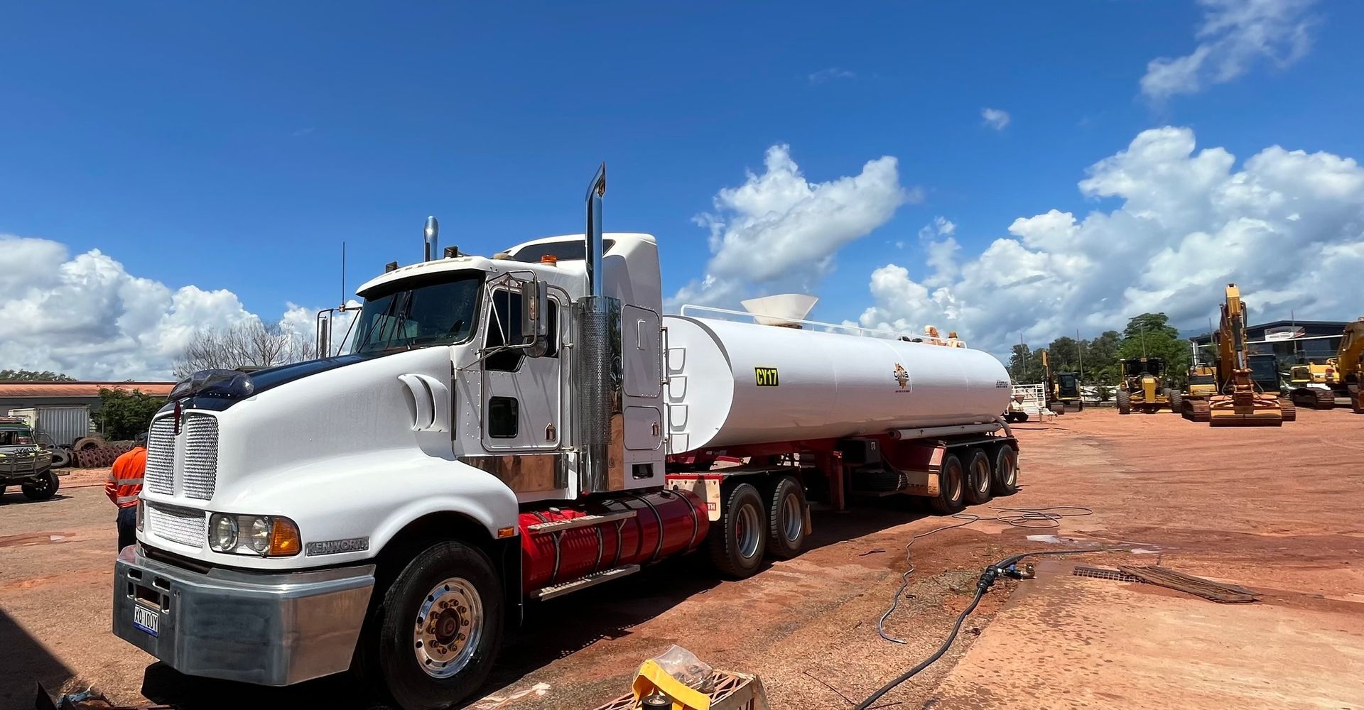 White tanker truck parked on a red dirt site under a blue sky with clouds, near construction equipment