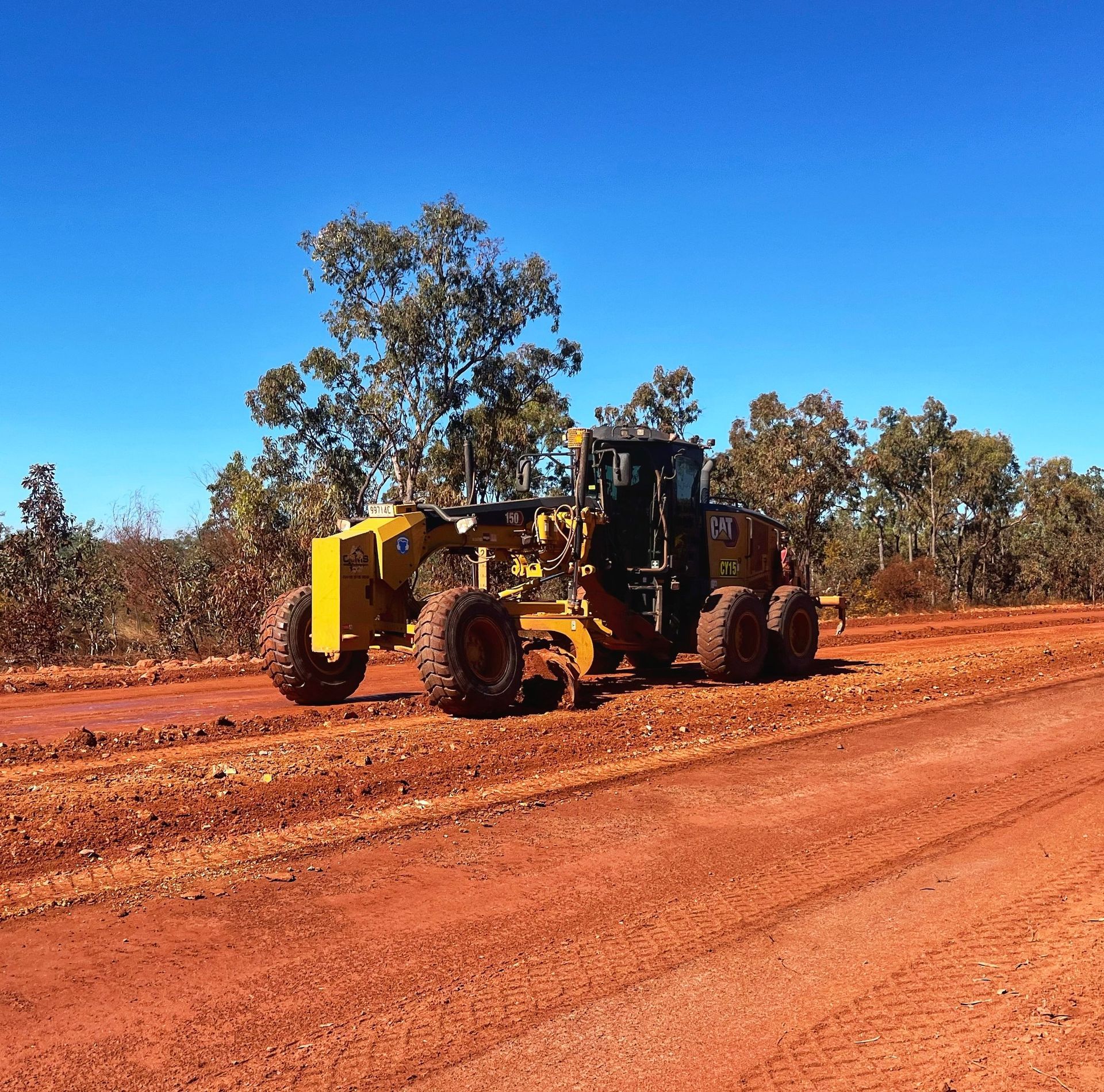 Yellow road grader on a red dirt road under a clear blue sky, with trees in the background
