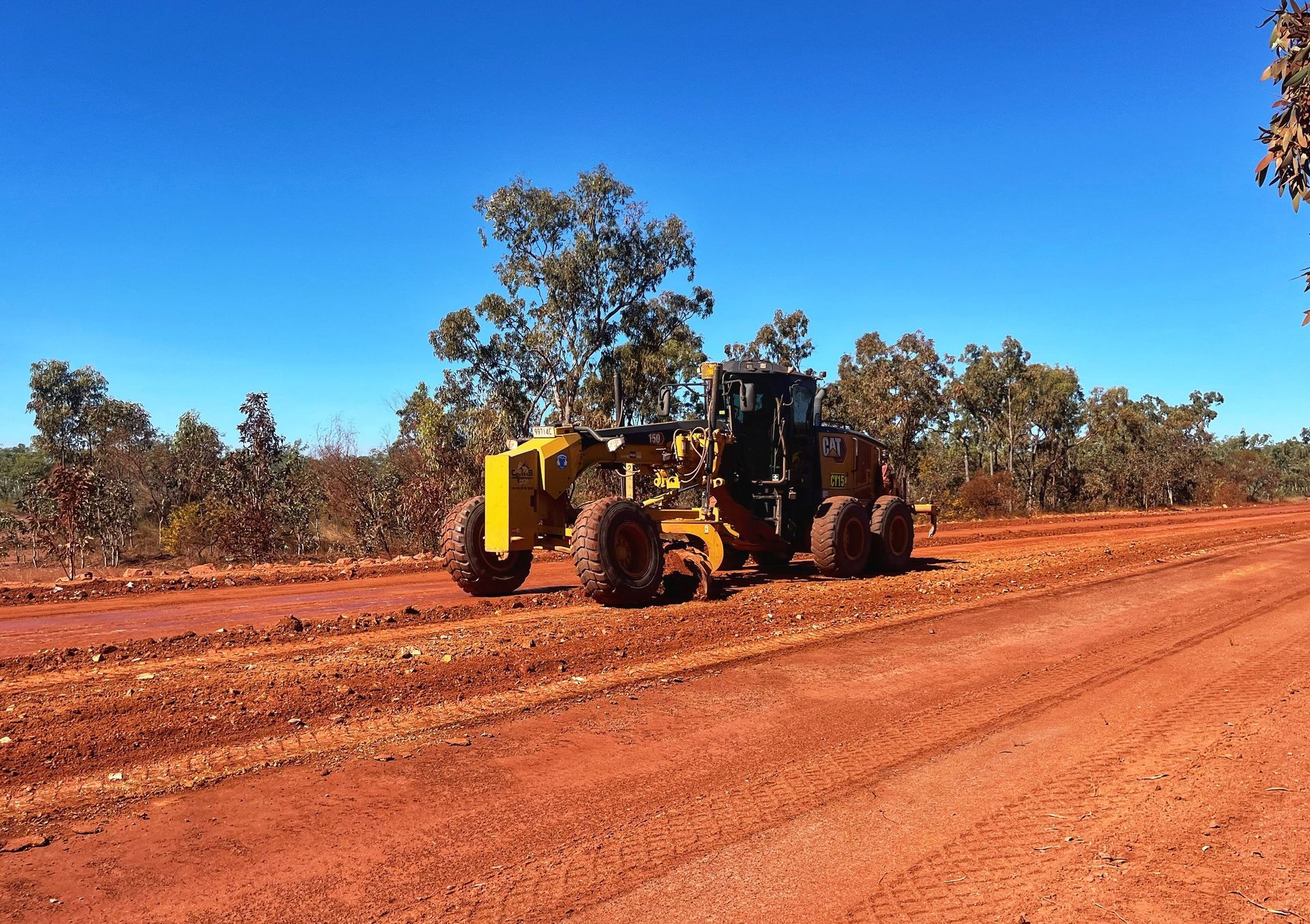 Yellow road grader on a red dirt road under a clear blue sky.