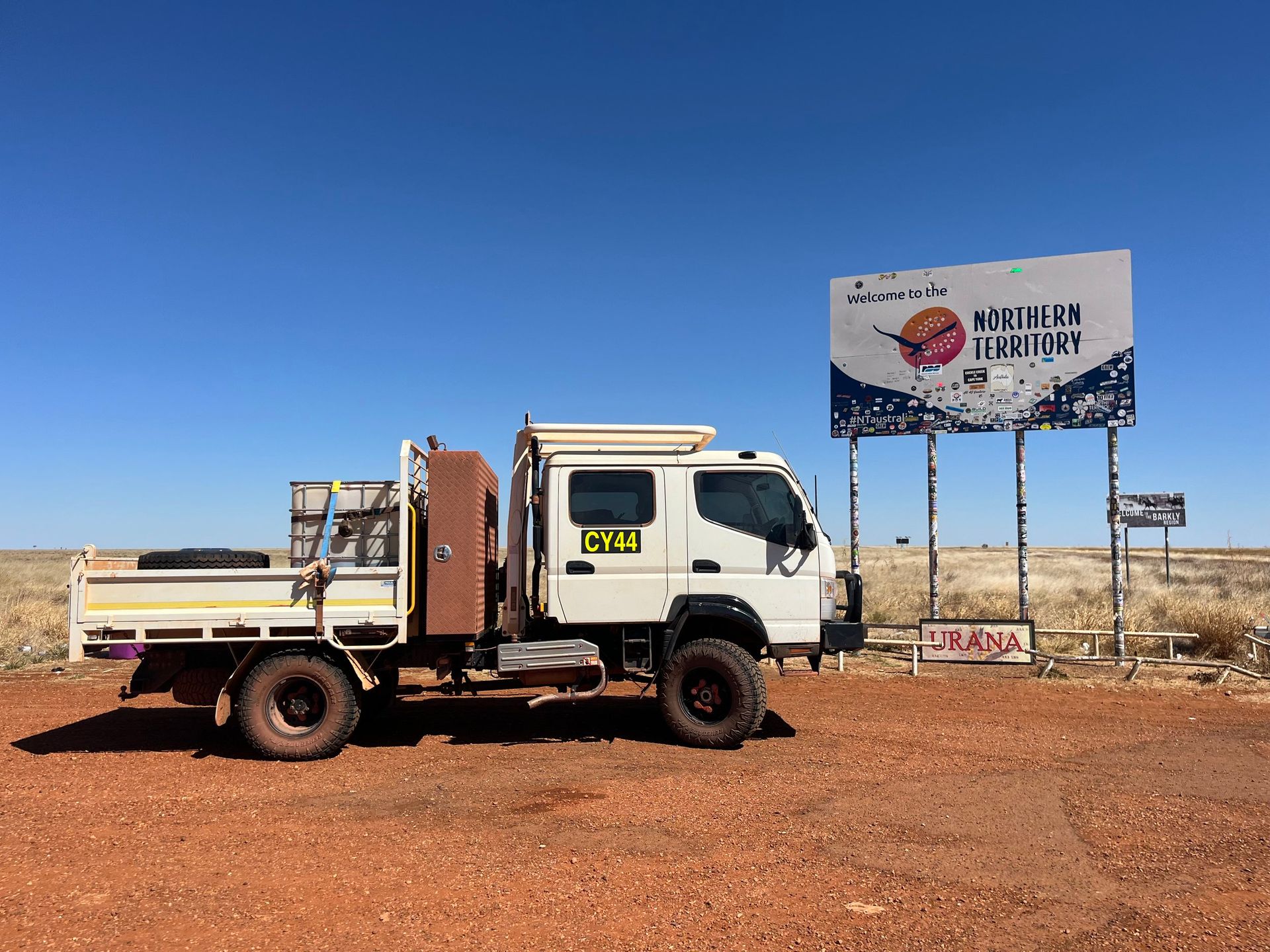 White flatbed truck parked on red dirt beside a roadside billboard under a clear blue sky.