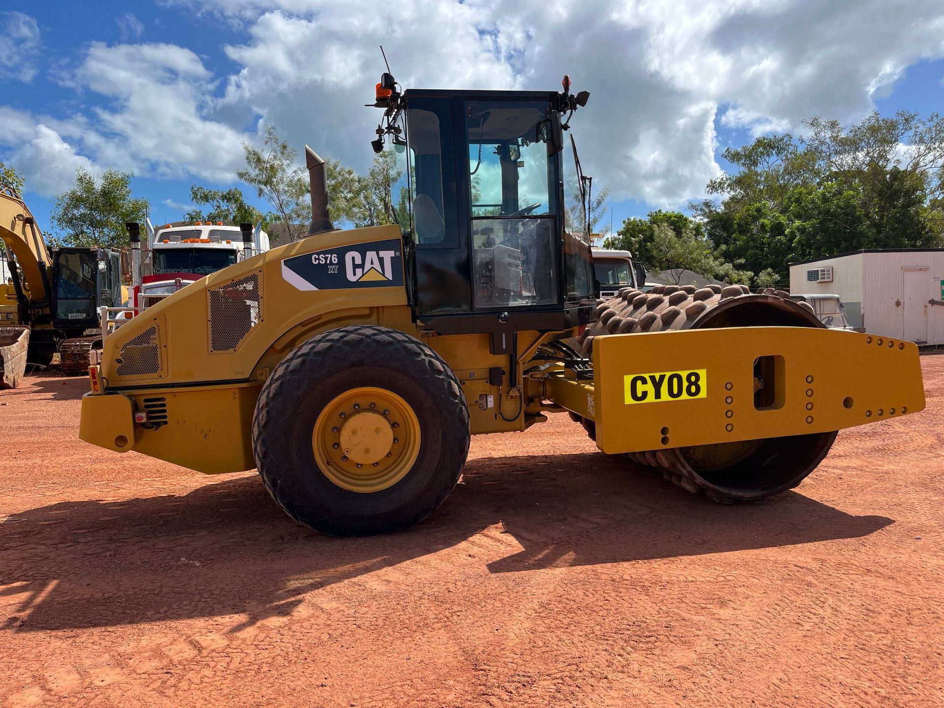 Yellow CAT road roller parked on red dirt under a partly cloudy sky