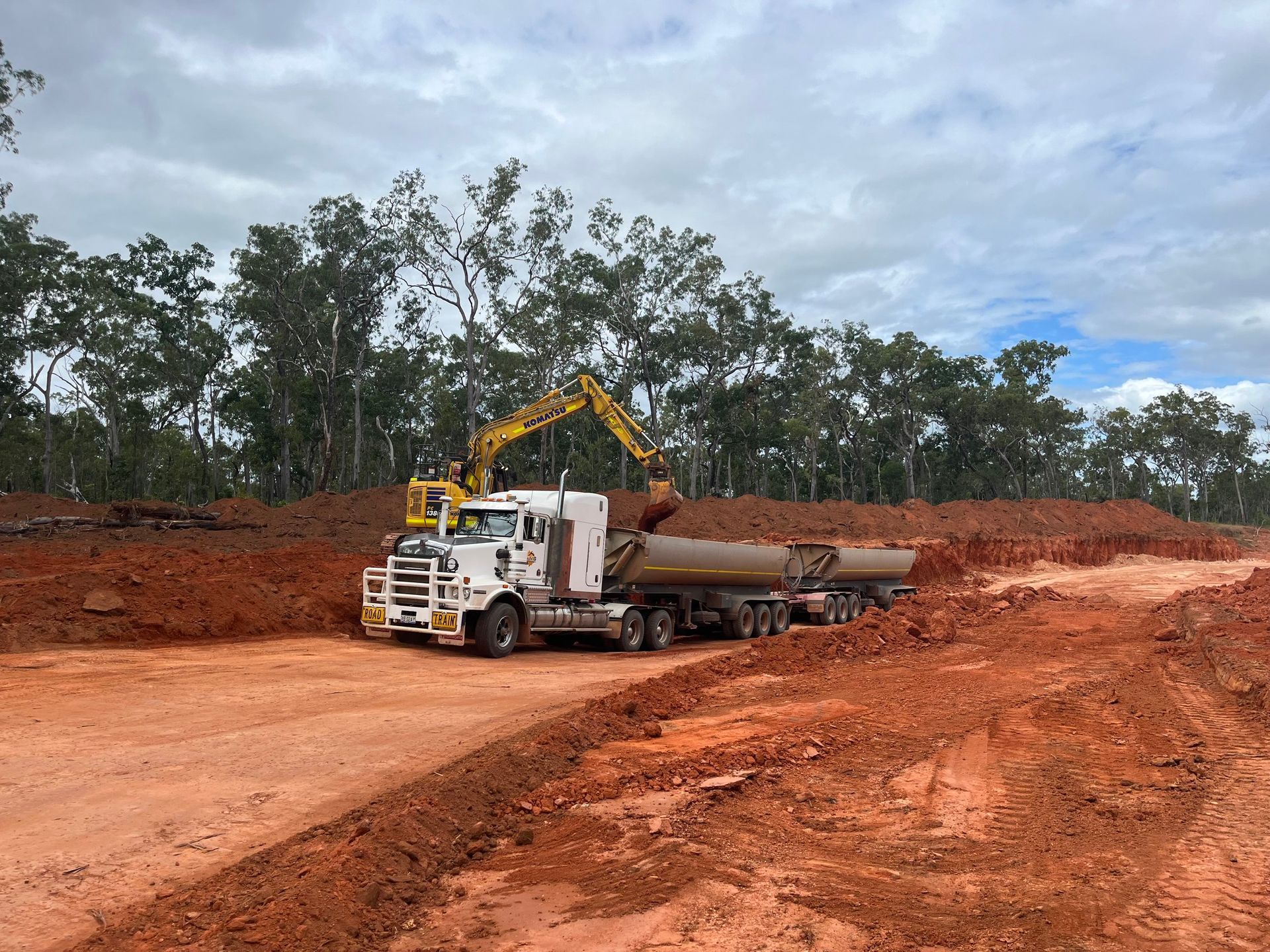 White dump truck and yellow excavator working on a red dirt road in a cleared wooded site.