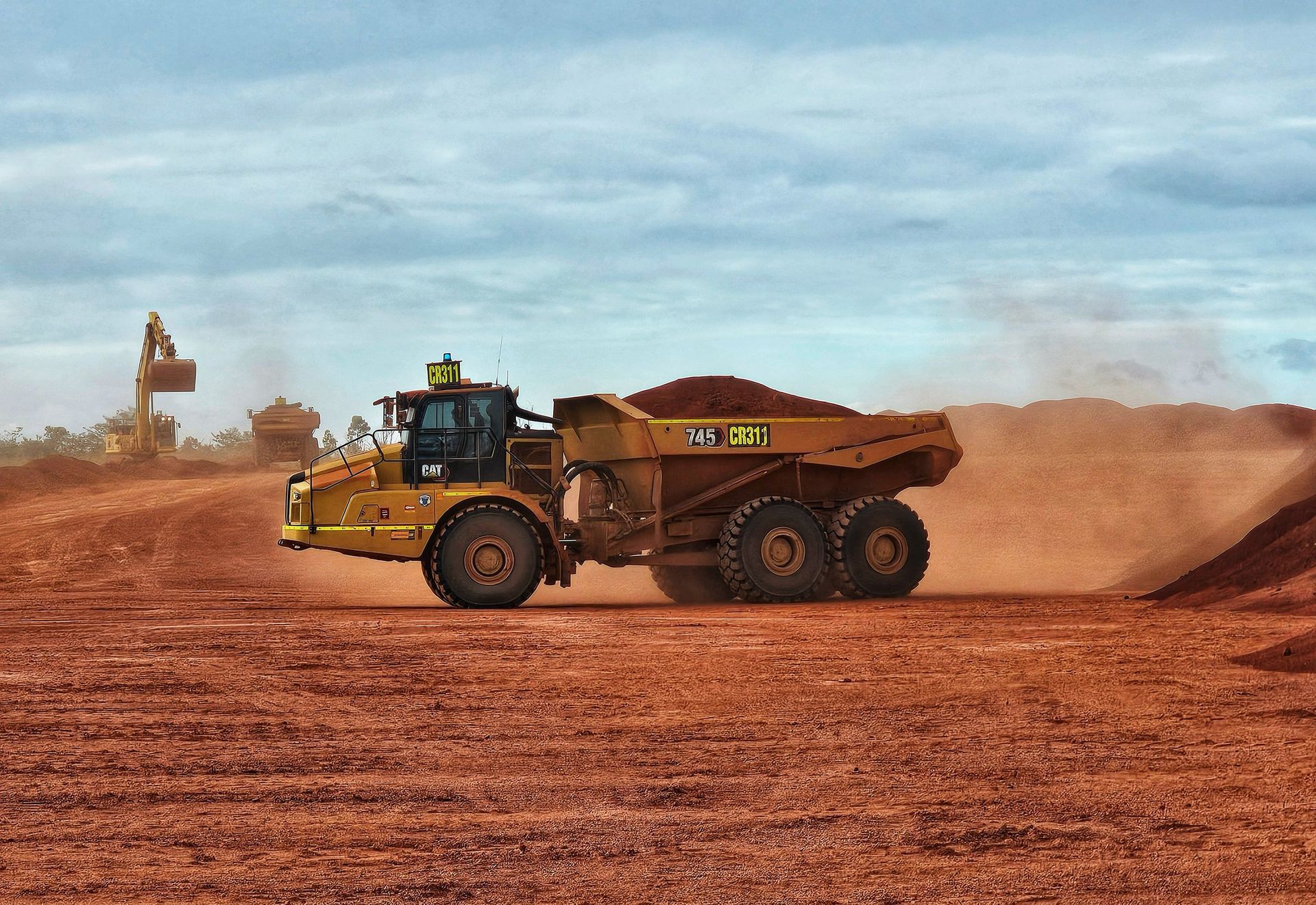 Yellow dump truck hauling dirt across a red dusty construction site under a cloudy sky