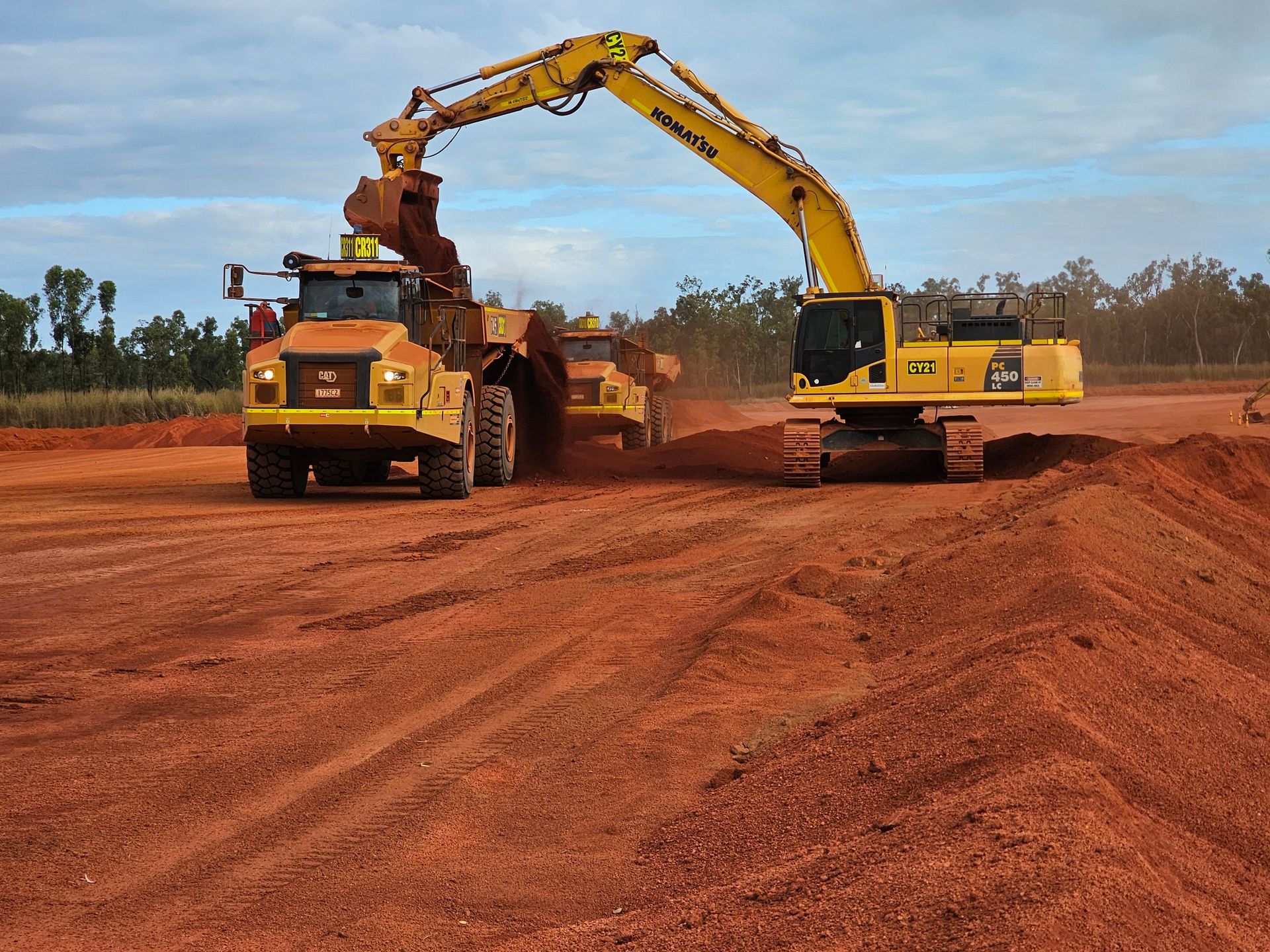 Yellow excavator loading red dirt into a dump truck at a construction site under a cloudy sky
