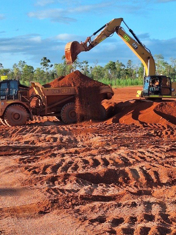 Excavator loading red dirt into a dump truck at a muddy construction site under a blue sky