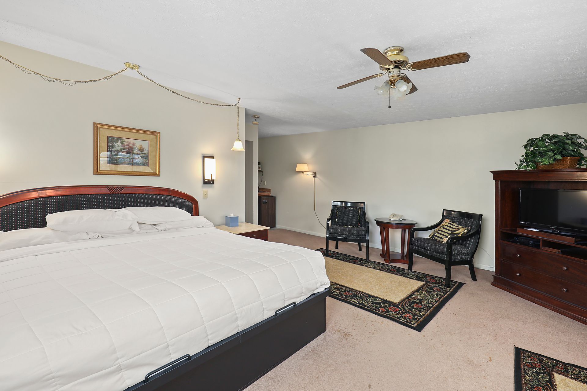 A bedroom with a king size bed , chairs , a television and a ceiling fan.