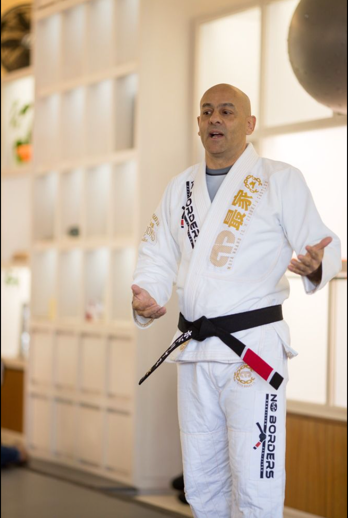 Man in white gi with black belt gestures while speaking, indoor setting.