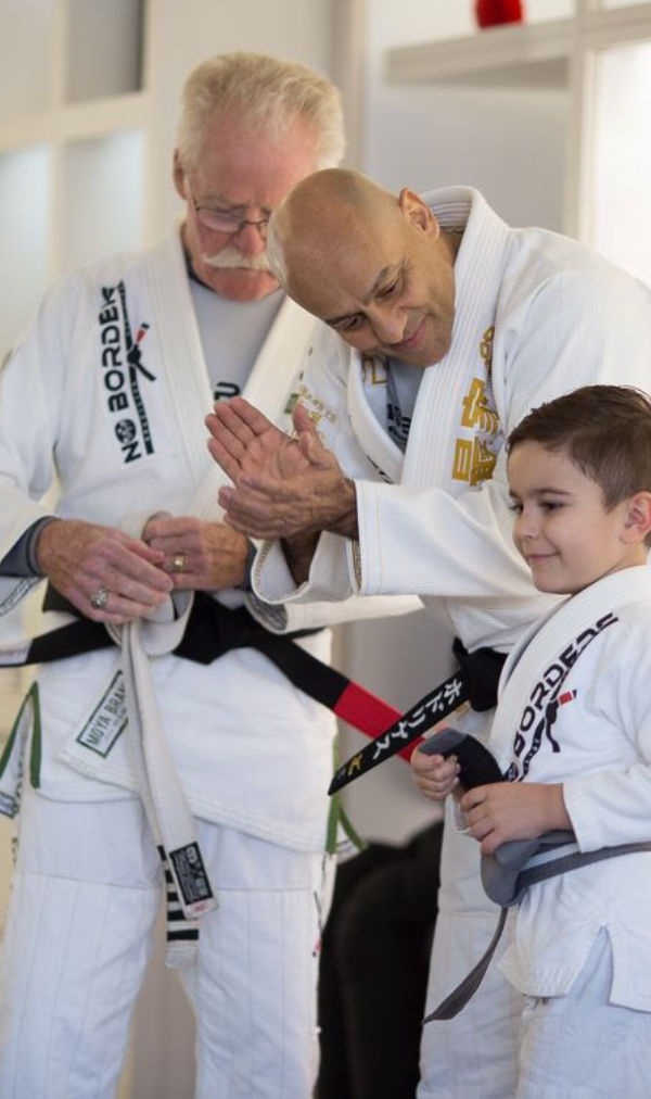 Three people tying a belt. One older man with white hair and a mustache, a middle-aged man and a child in a martial arts gi.