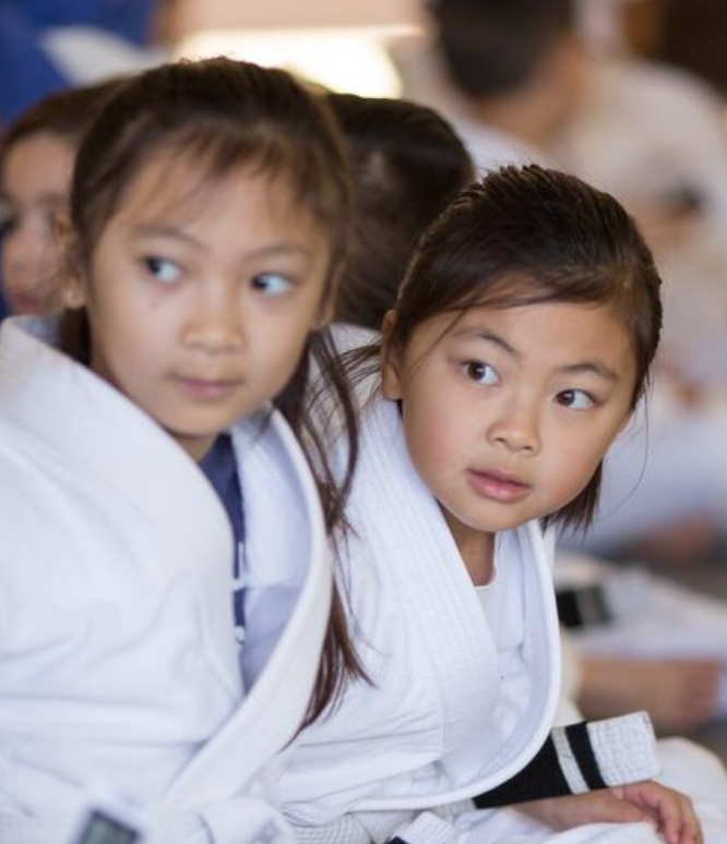 Two young girls in white martial arts uniforms look on attentively.