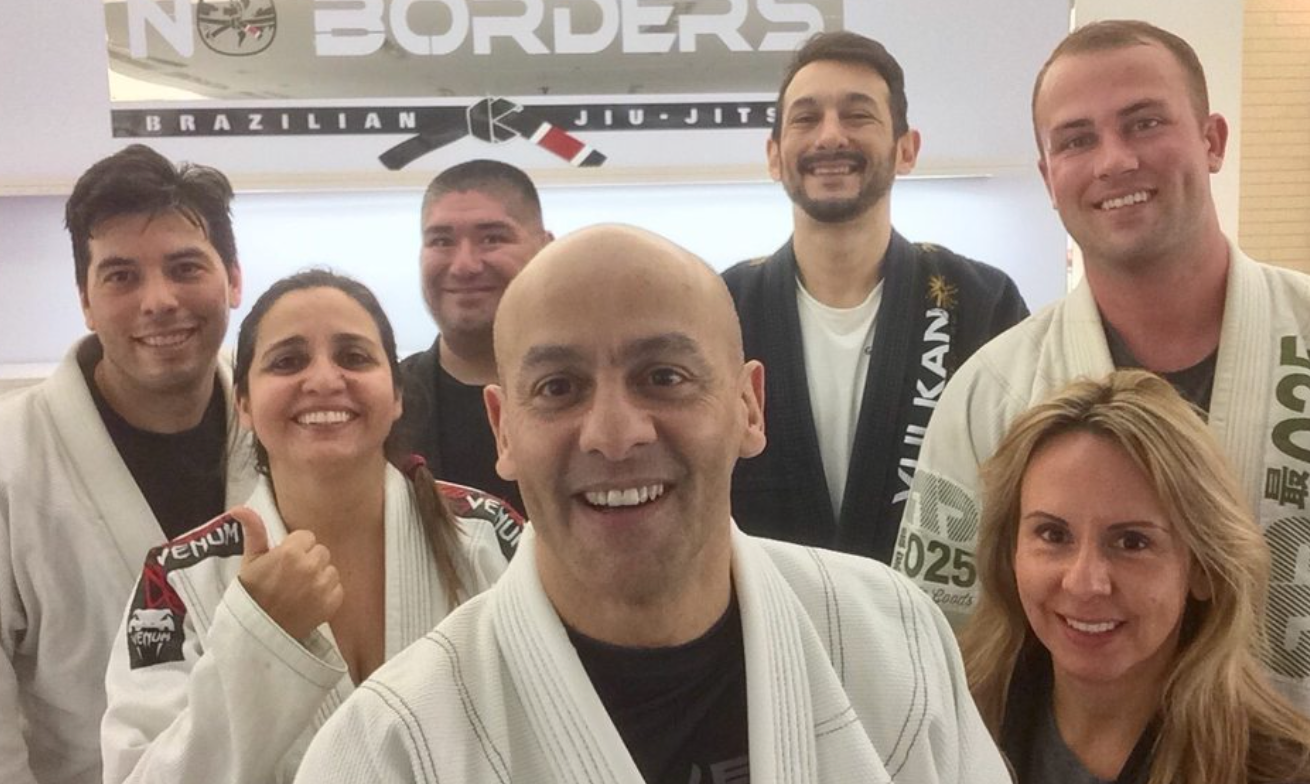 Group of smiling people in martial arts uniforms, standing in front of a Brazilian Jiu-Jitsu gym, light-skinned and dark-haired.