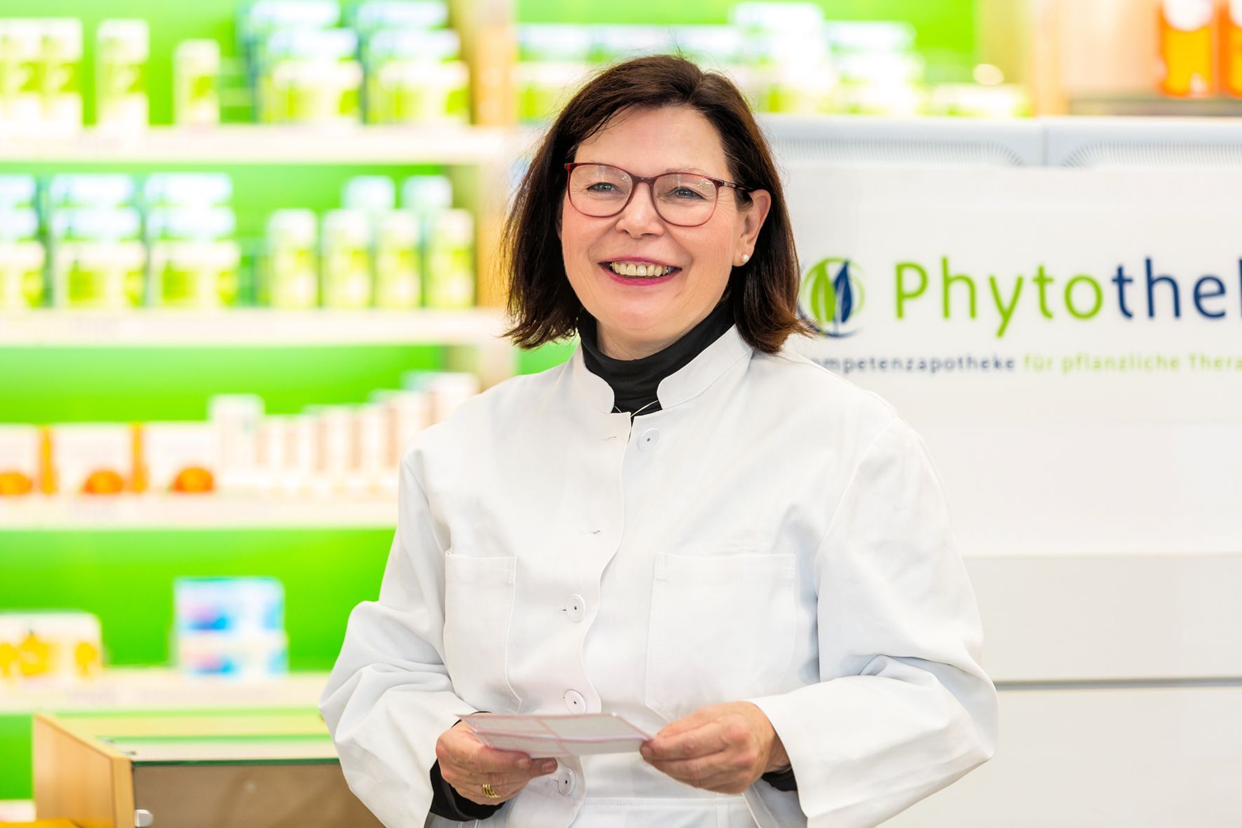 Eine Frau in einem weißen Laborkittel und mit Brille steht in einer Apotheke.