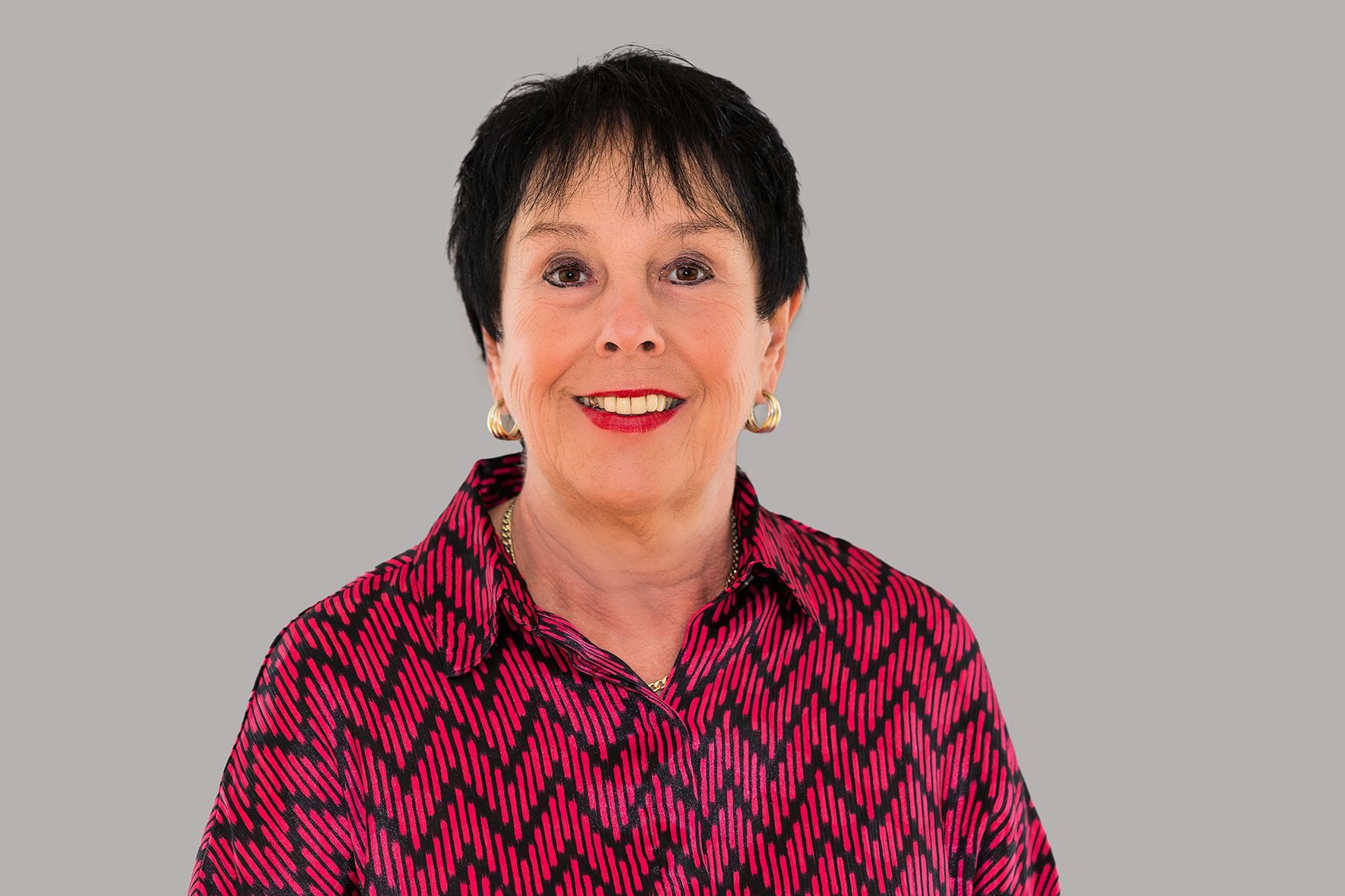 Eine Frau in einem rosa Hemd und mit Ohrringen lächelt in die Kamera.