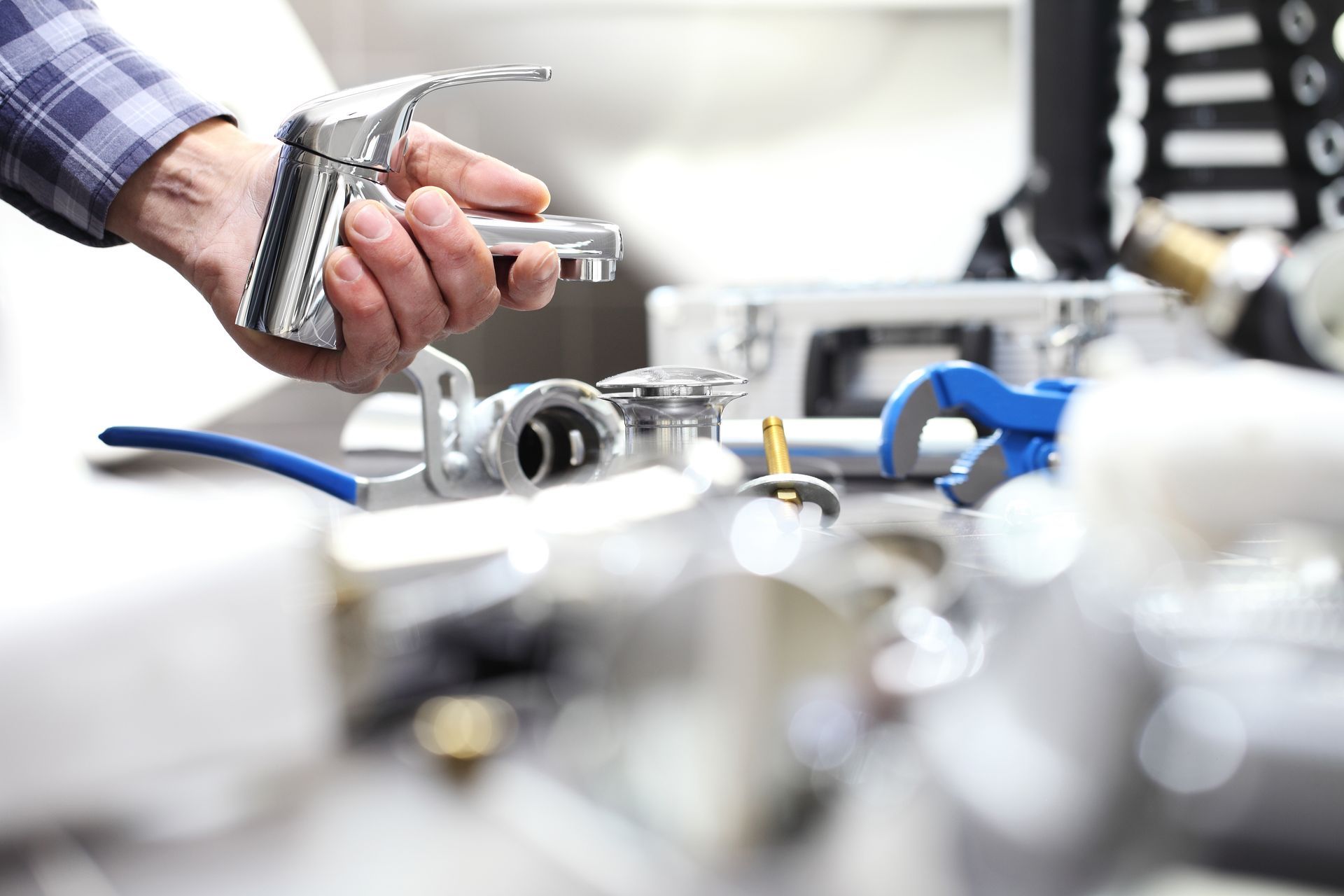 A hand holds a chrome bathroom faucet above a workbench with plumbing tools and parts.