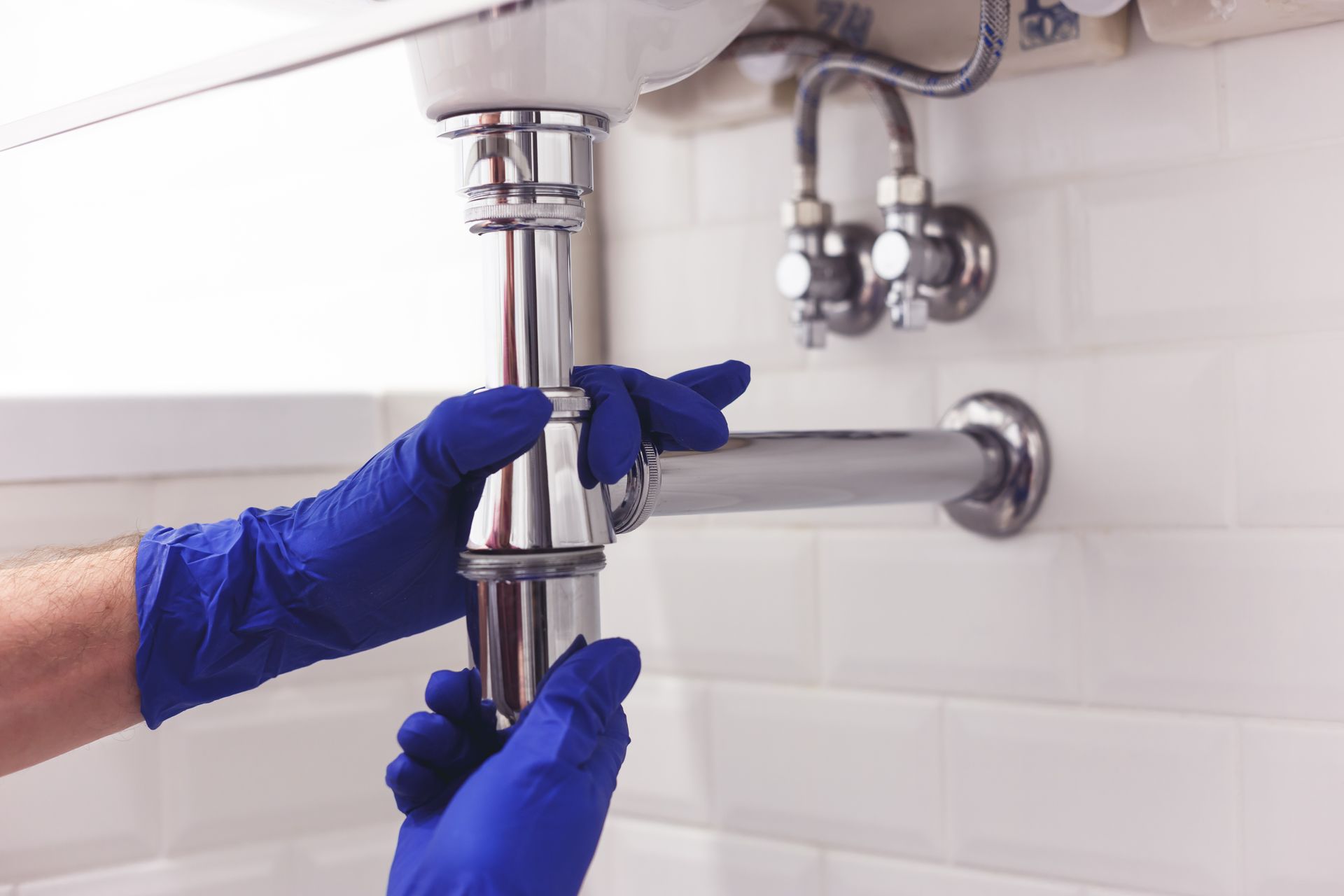 A pair of hands in blue gloves tightening a chrome sink drain pipe assembly against a white tiled wall.