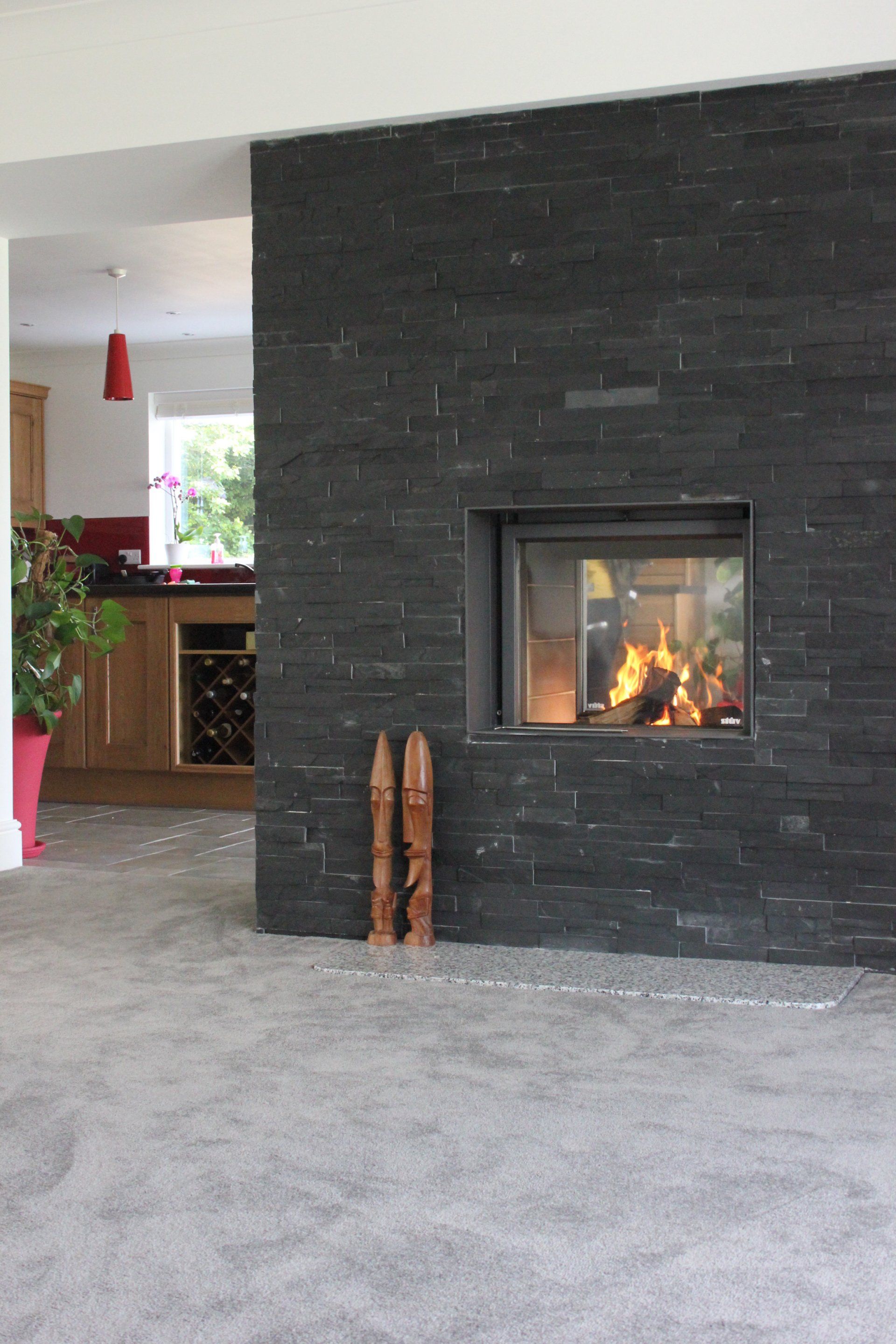 Fireplace with lit fire in a dark brick wall, opening into a kitchen with light wood cabinets.