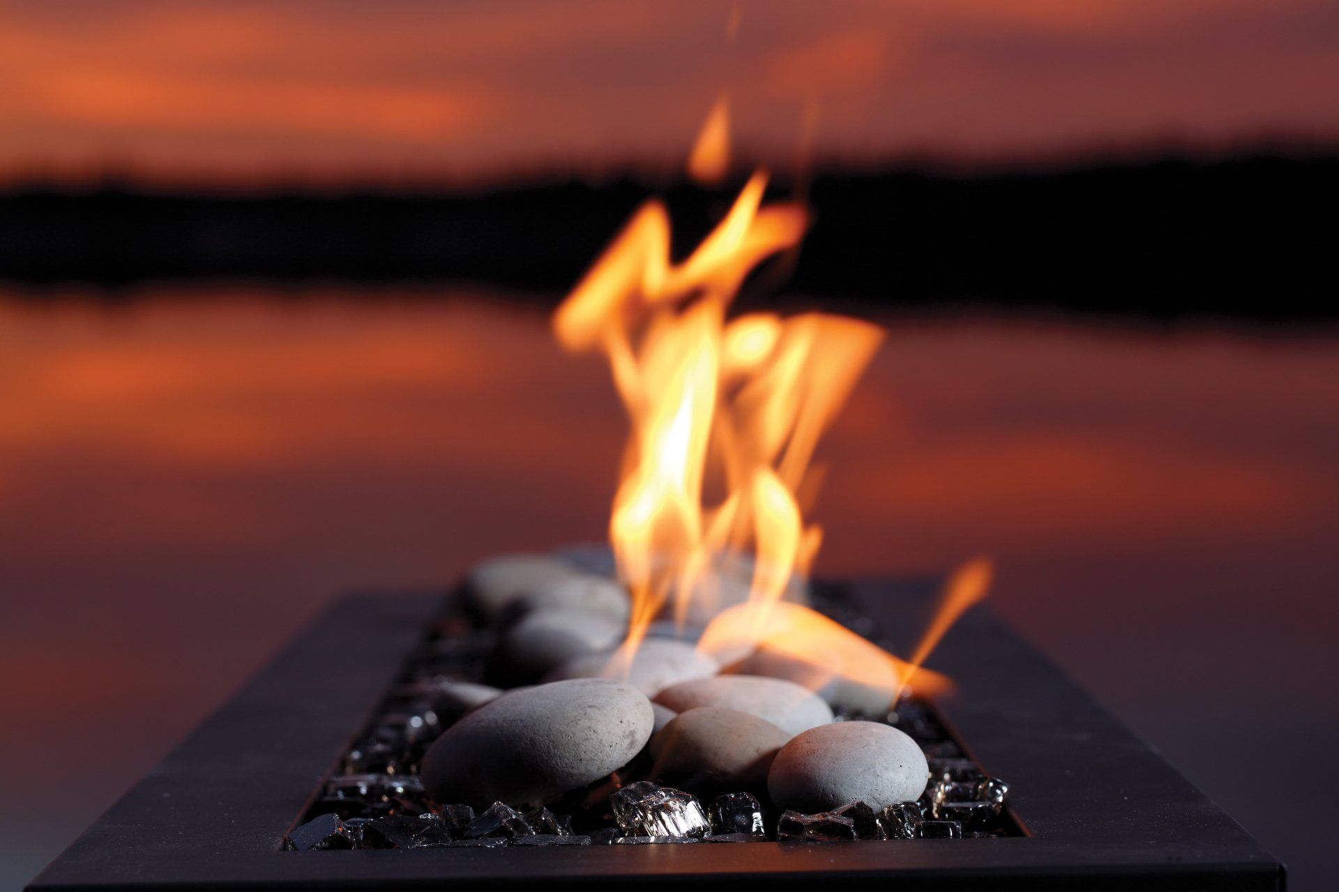 Fire pit with flames, gray river rocks, black base, and sunset background.