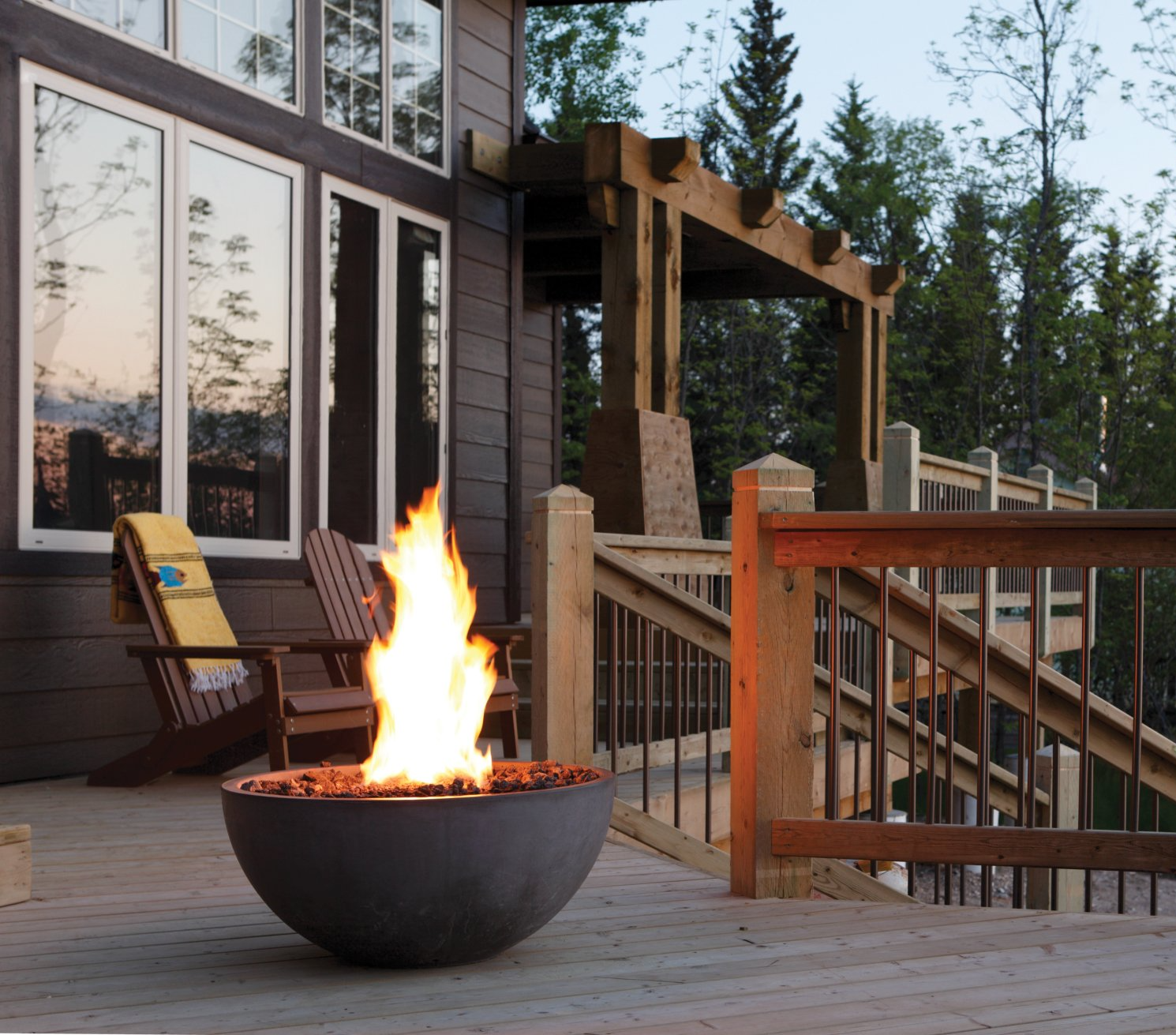 Fire bowl with flames on a wooden deck, near a house with large windows and a pergola.