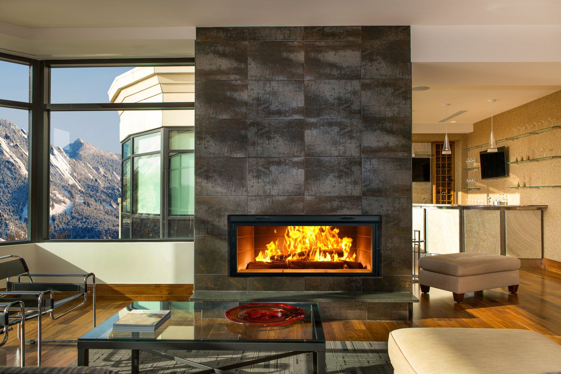 Modern fireplace with burning flames, built into a dark stone wall, with mountain view outside a large window.