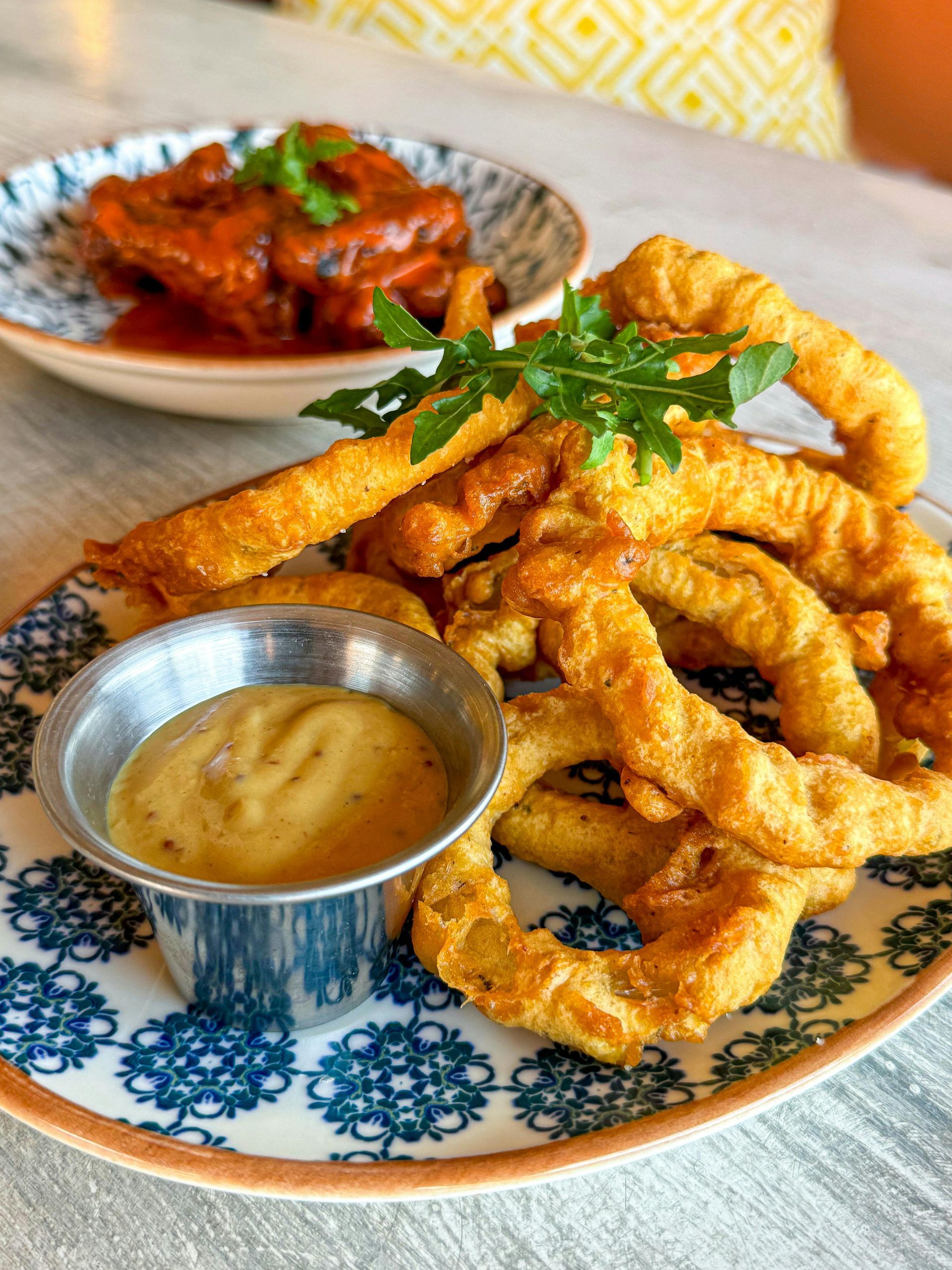 Fried calamari and sauce on a patterned plate, with red food in the background.