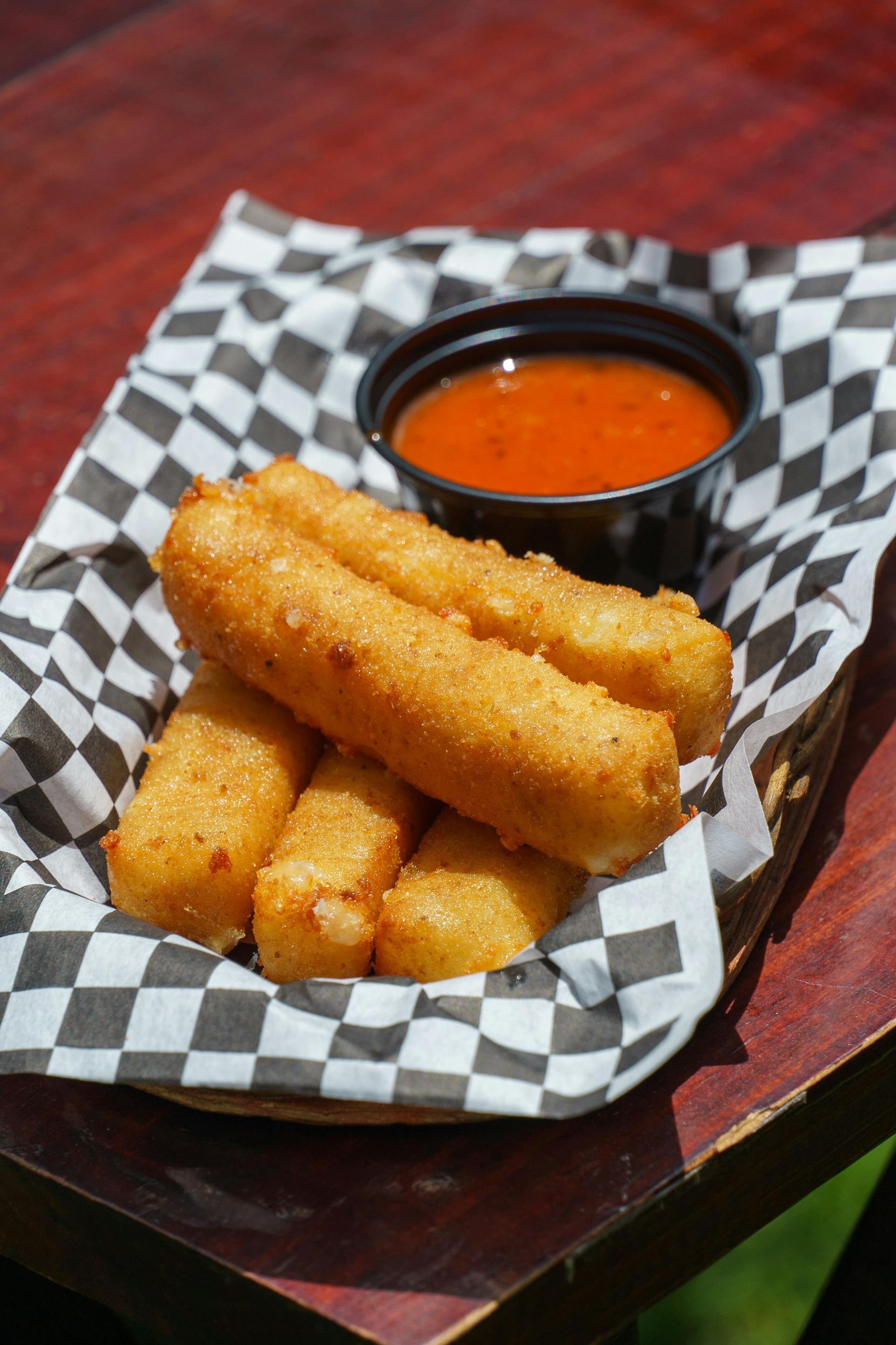 Golden fried mozzarella sticks with red dipping sauce in a basket lined with checkered paper.